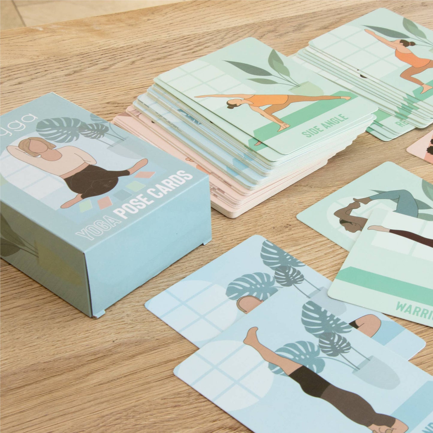 The retail box for the Myga Yoga Pose Cards sits on a wooden surface next to a full stack of instructional cards. Several individual cards are spread in the foreground, showing illustrations of various yoga poses like Side Angle and Warrior variations, demonstrating the set's contents.