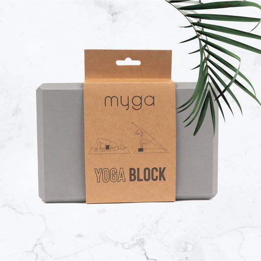 The Myga grey foam Yoga Block is displayed with its kraft paper retail packaging wrapped around it. The packaging shows illustrations of how the block is used and is labeled 'YOGA BLOCK'. The product is set on a marble background with a palm leaf visible on the side.