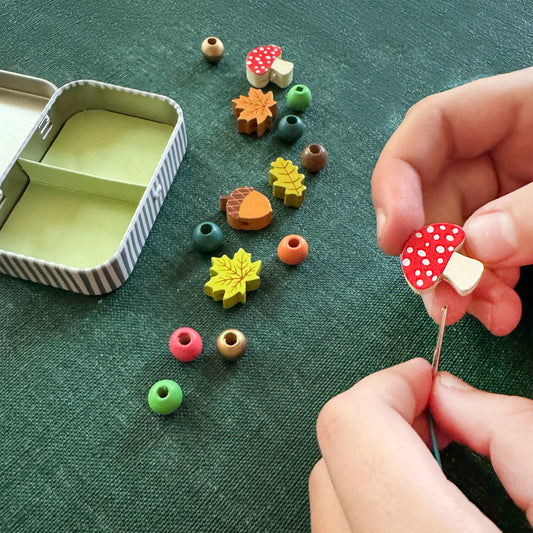 Close-up of a child's hands threading a red and white mushroom bead onto a needle and string. The wooden beads are spread on a green surface, showing a variety of autumn-themed charms including maple leaves and acorns, next to the open, empty storage tin from the 'Forest Bracelet Kit.'
