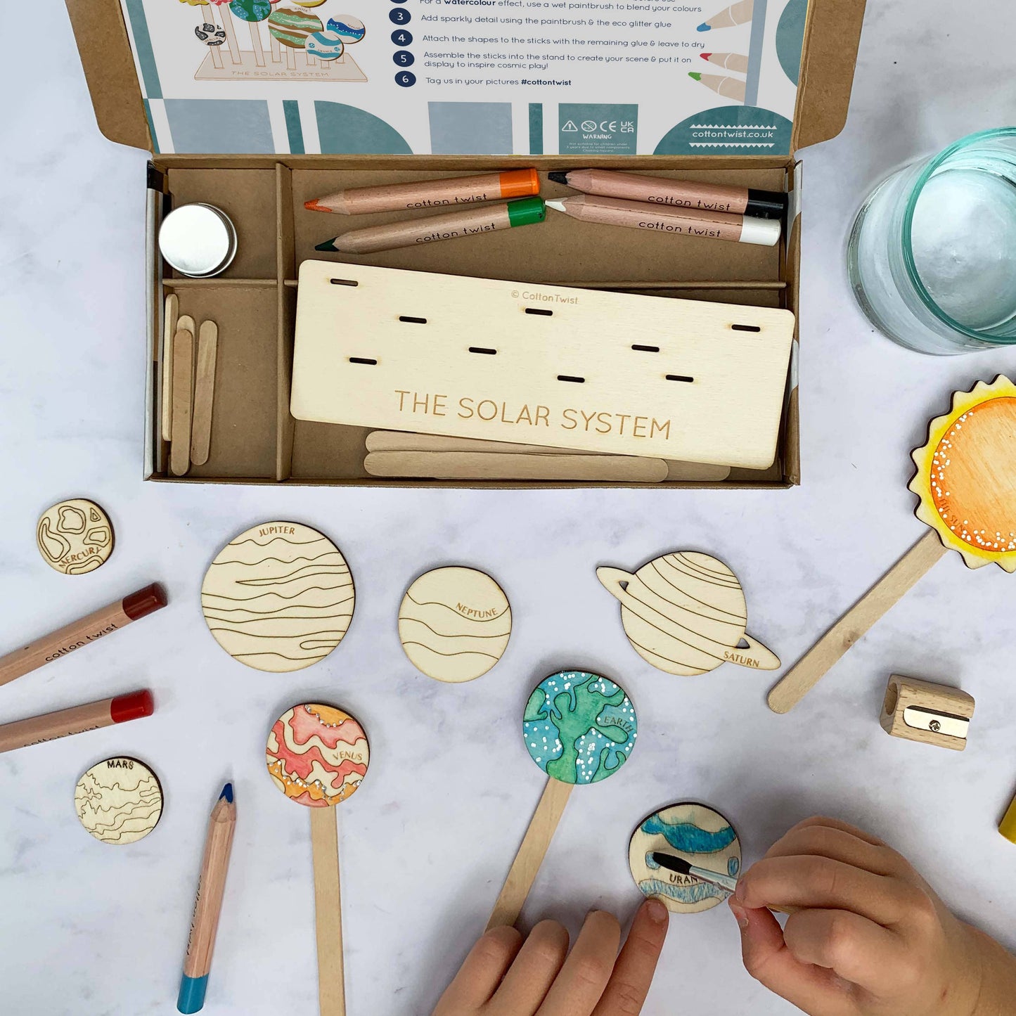 A child's hands work on the DIY Solar System craft kit, coloring or attaching the wooden planet tokens onto the sticks. The open kit box and wooden display stand are visible in the background, along with the pencils and blank wooden planet cutouts spread on a marble-style table