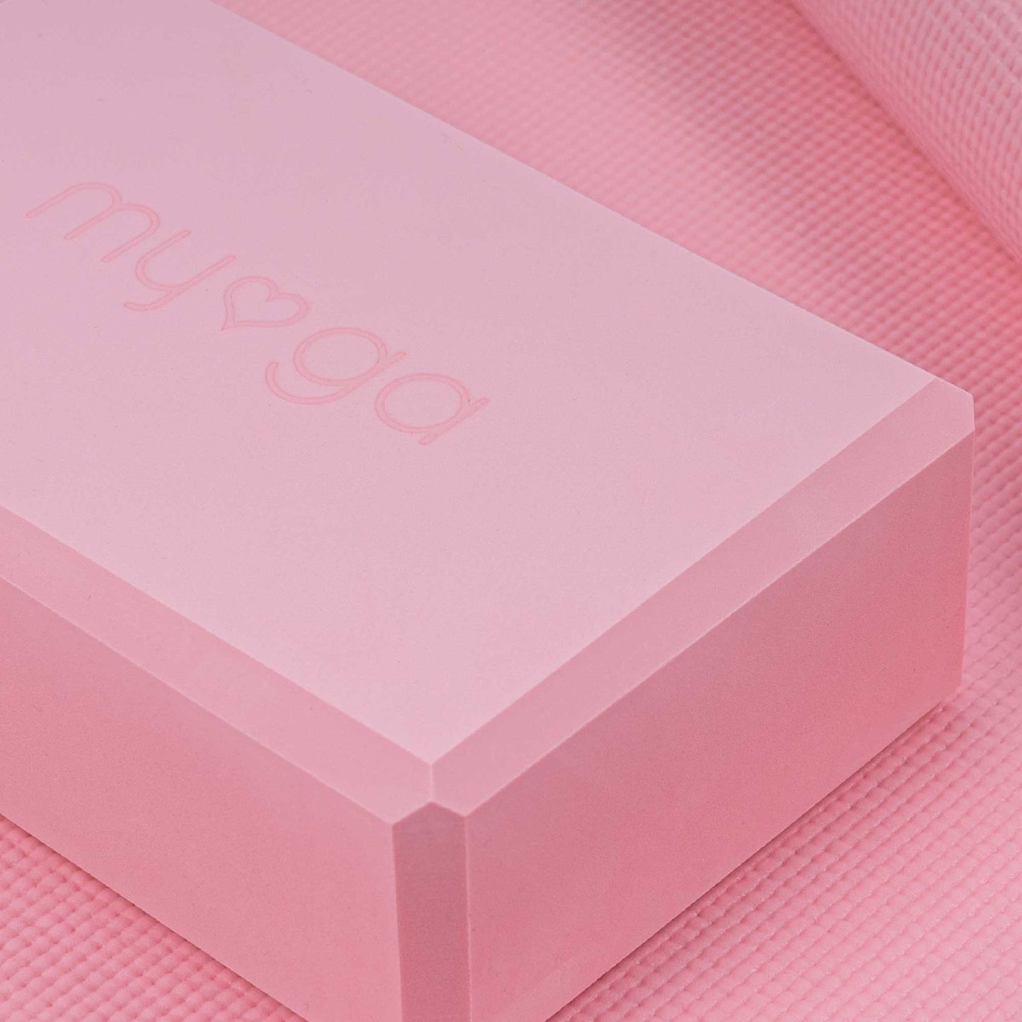 Extreme close-up of a pink Myga foam yoga block resting on a matching pink yoga mat. The block features the embossed Myga logo, highlighting the product's color and texture.