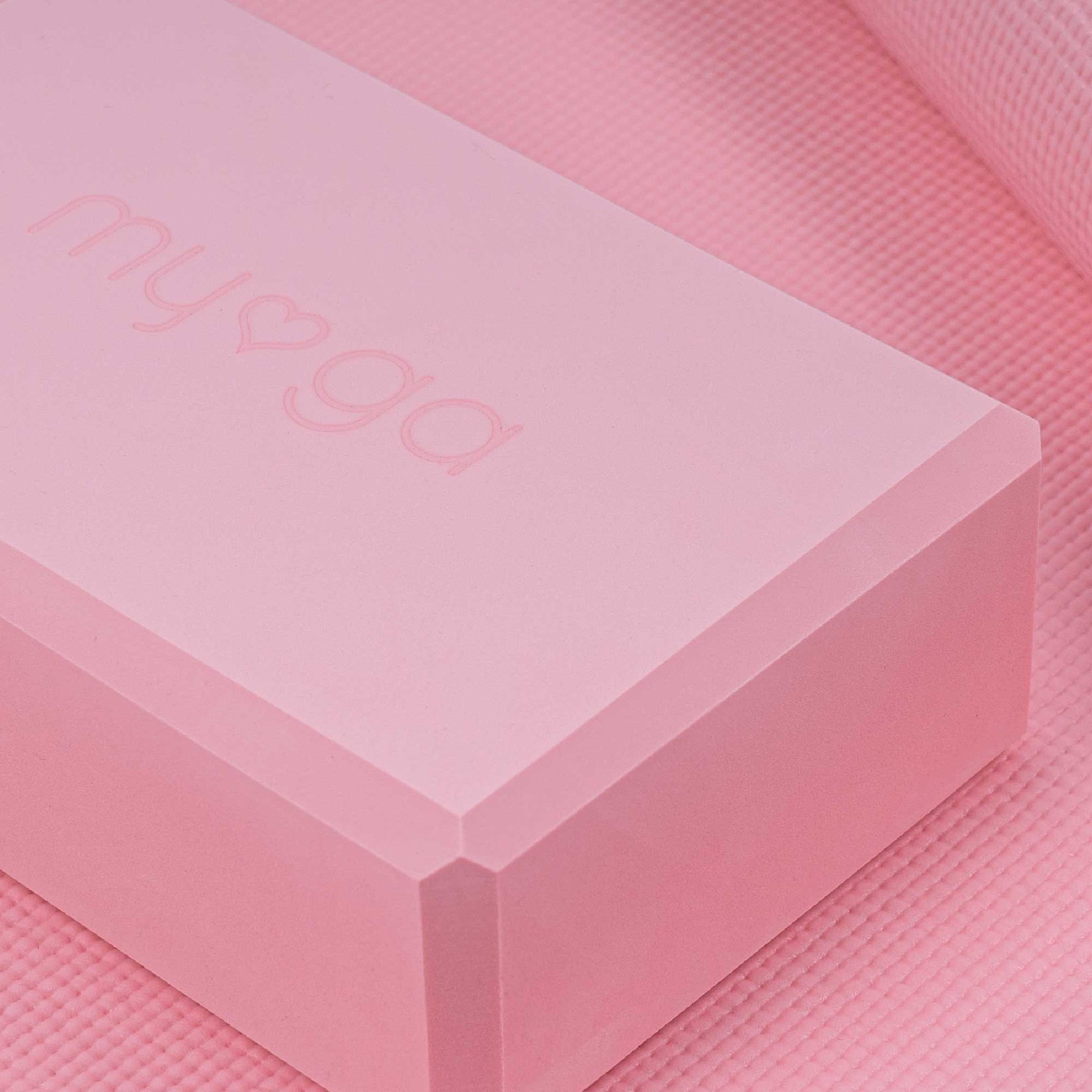 Extreme close-up of a pink Myga foam yoga block resting on a matching pink yoga mat. The block features the embossed Myga logo, highlighting the product's color and texture.