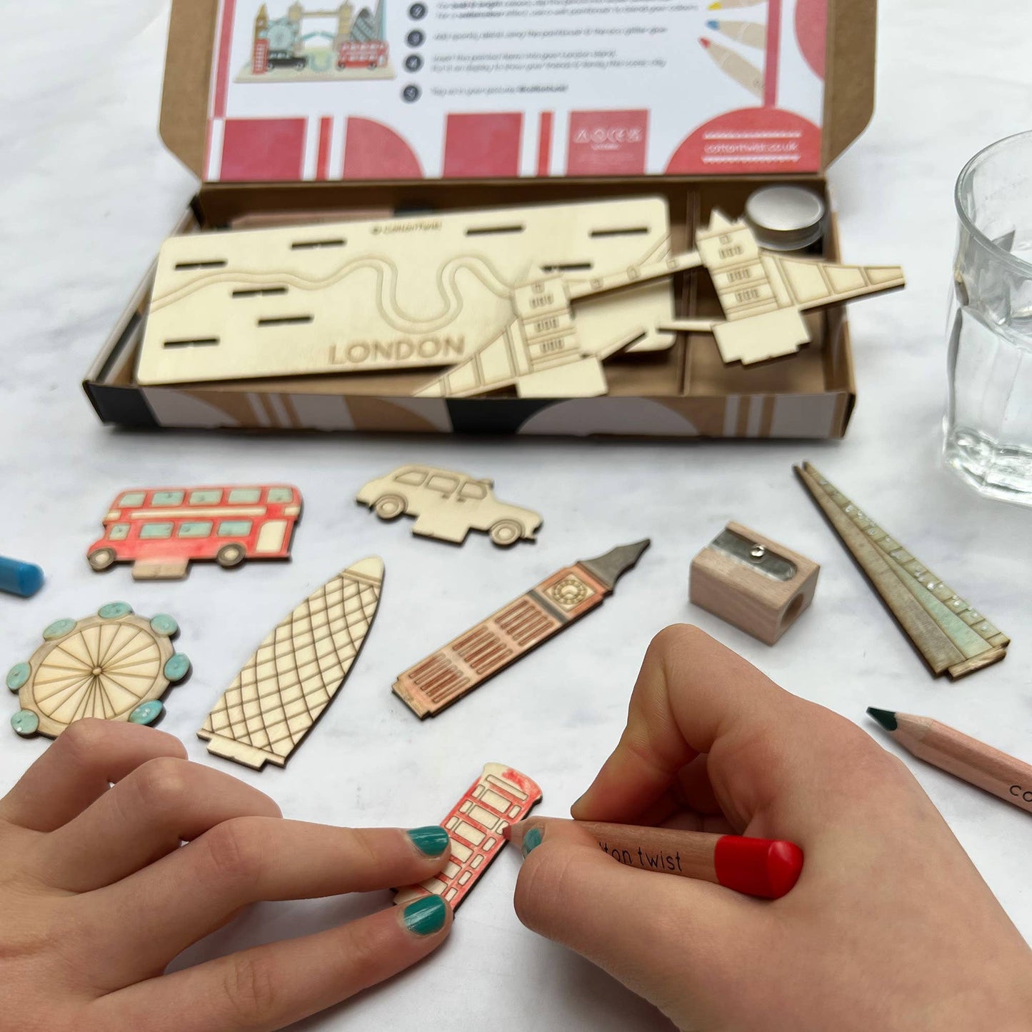 A child's hands are coloring the red double-decker bus wooden piece with a red pencil from the DIY London Landmarks craft kit. The components, including wooden pieces for Big Ben, a taxi, the London Eye, and the base labeled 'LONDON,' are spread out, showing the coloring activity in progress.