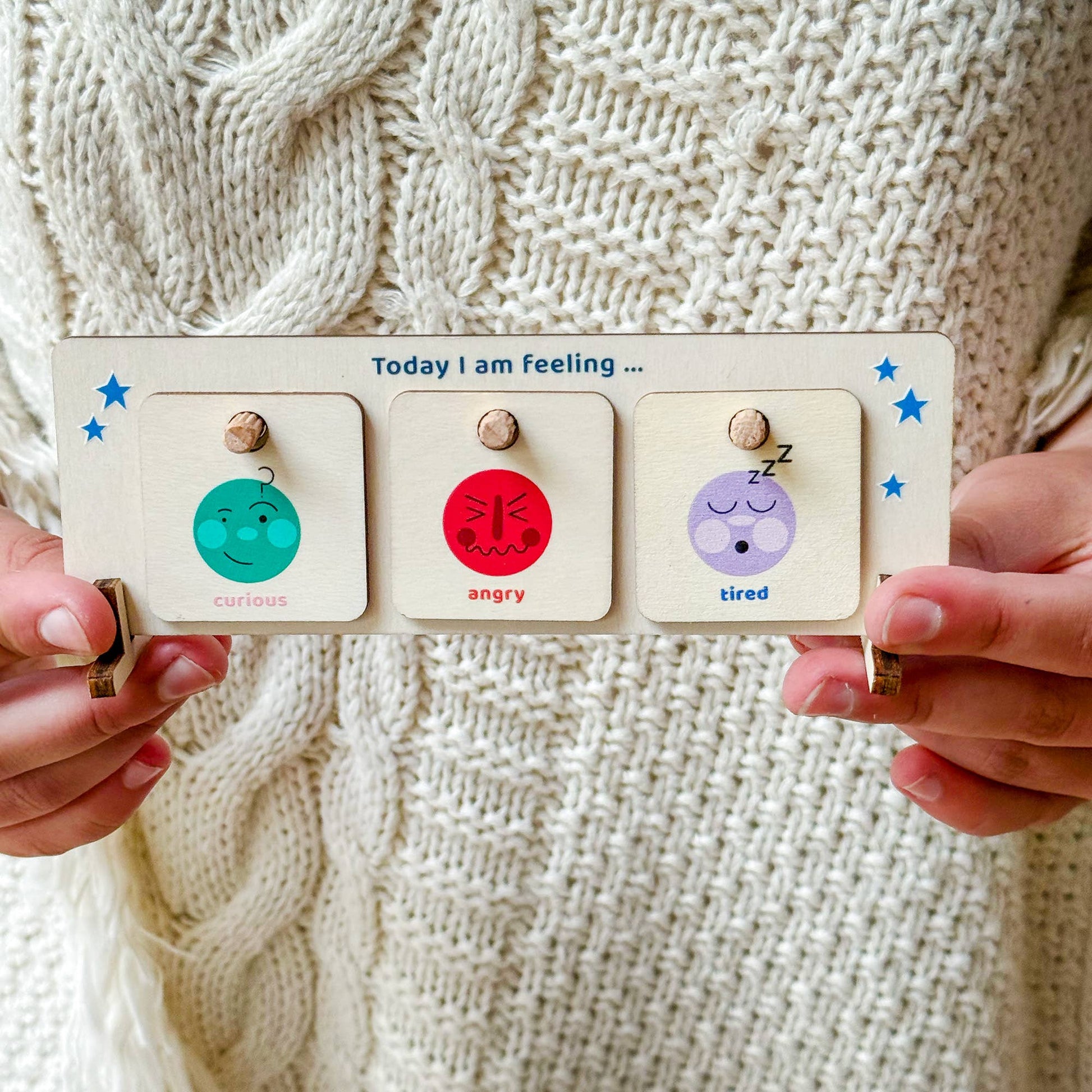 Close-up of a wooden "Today I am feeling" chart held by someone wearing a white knitted sweater. The chart displays three emotion tiles: 'curious,' 'angry,' and 'tired.' The educational mood board is a social-emotional learning tool for children.