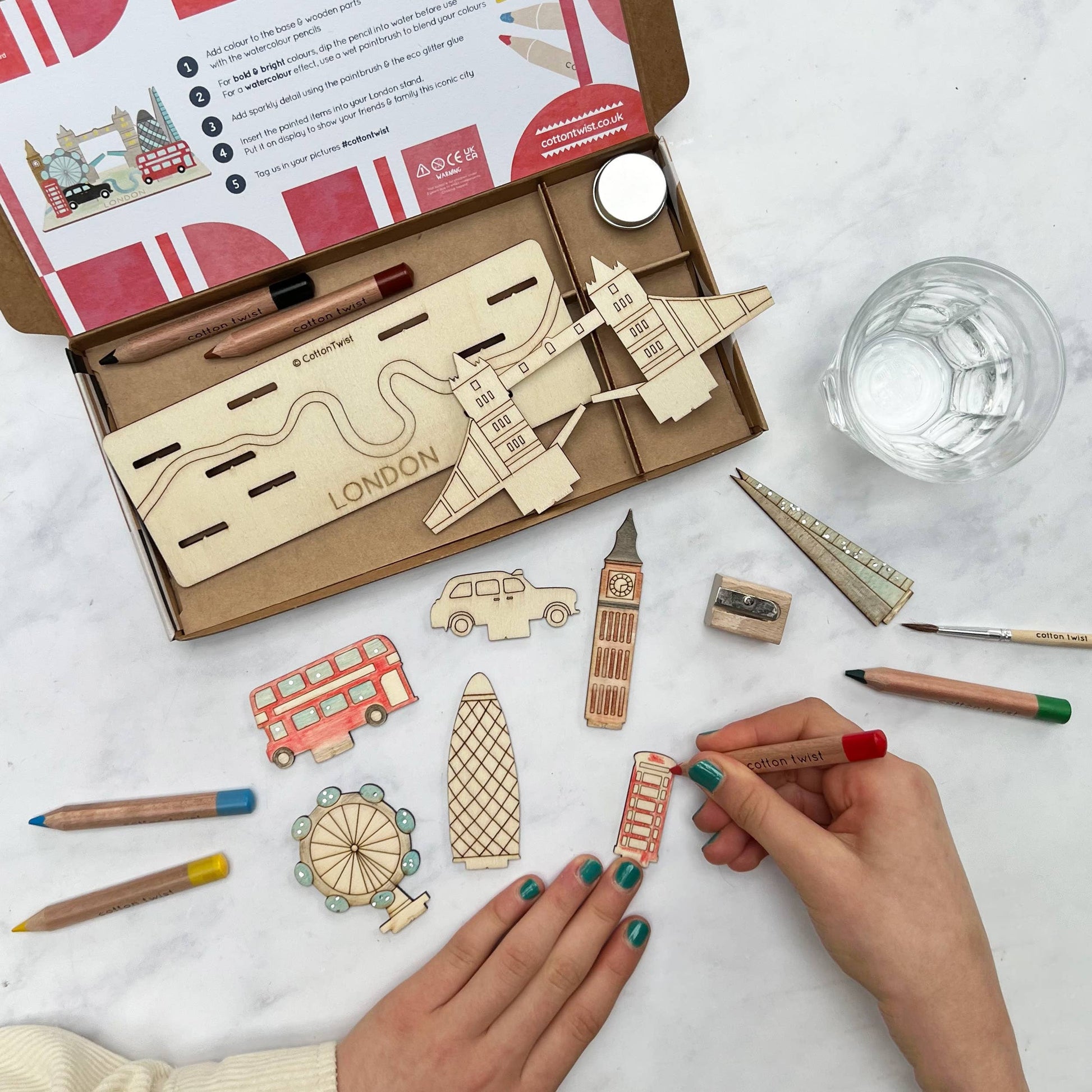 Overhead view of a child's hands coloring a wooden London telephone booth piece with a red pencil from the DIY London Landmarks craft kit. The open box contains the unpainted 3D model base labeled 'LONDON.' Pieces like Big Ben, the double-decker bus, and the London Eye are spread around the work area, along with pencils and a paintbrush.