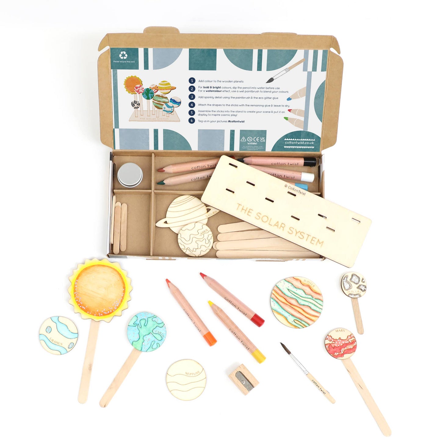 Complete contents of the DIY Solar System craft kit laid out, showing the open cardboard box organized with wooden planet tokens, a wooden stand, colored pencils, a pencil sharpener, a paintbrush, and other accessories. Several tokens have been partially or fully colored, demonstrating the craft activity.