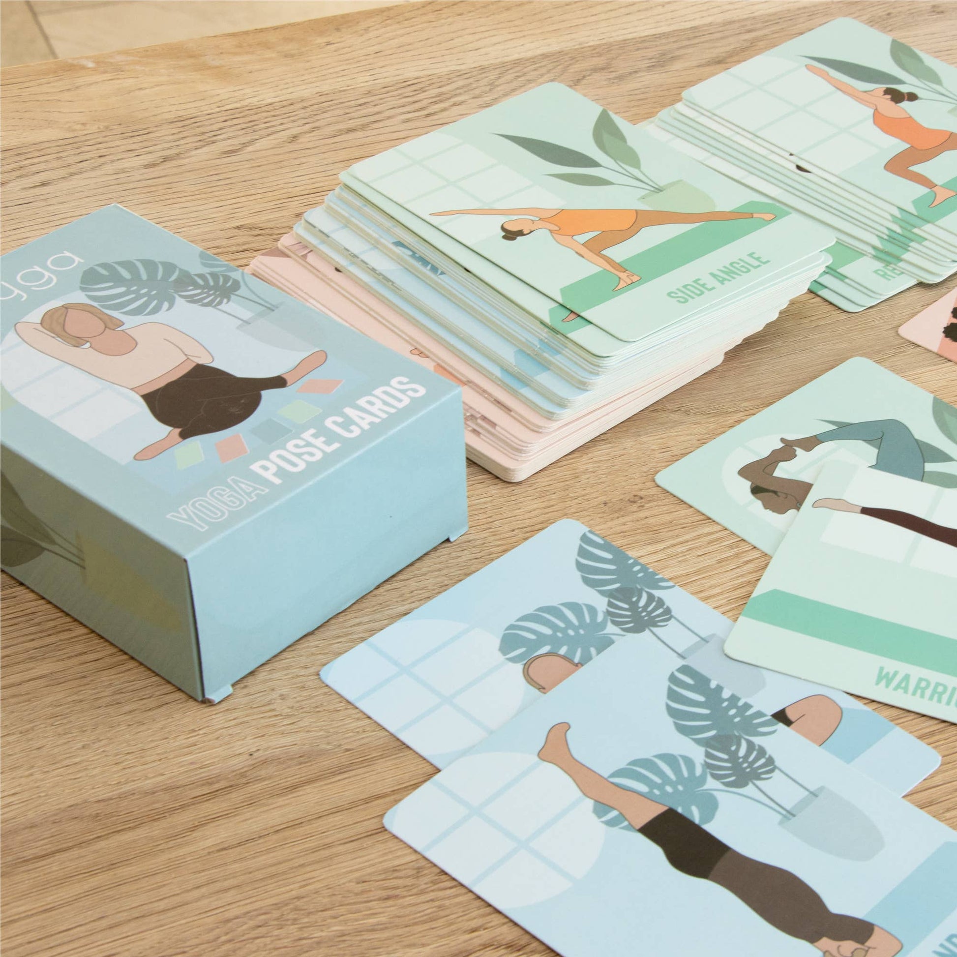 The retail box for the Myga Yoga Pose Cards sits on a wooden surface next to a full stack of instructional cards. Several individual cards are spread in the foreground, showing illustrations of various yoga poses like Side Angle and Warrior variations, demonstrating the set's contents.
