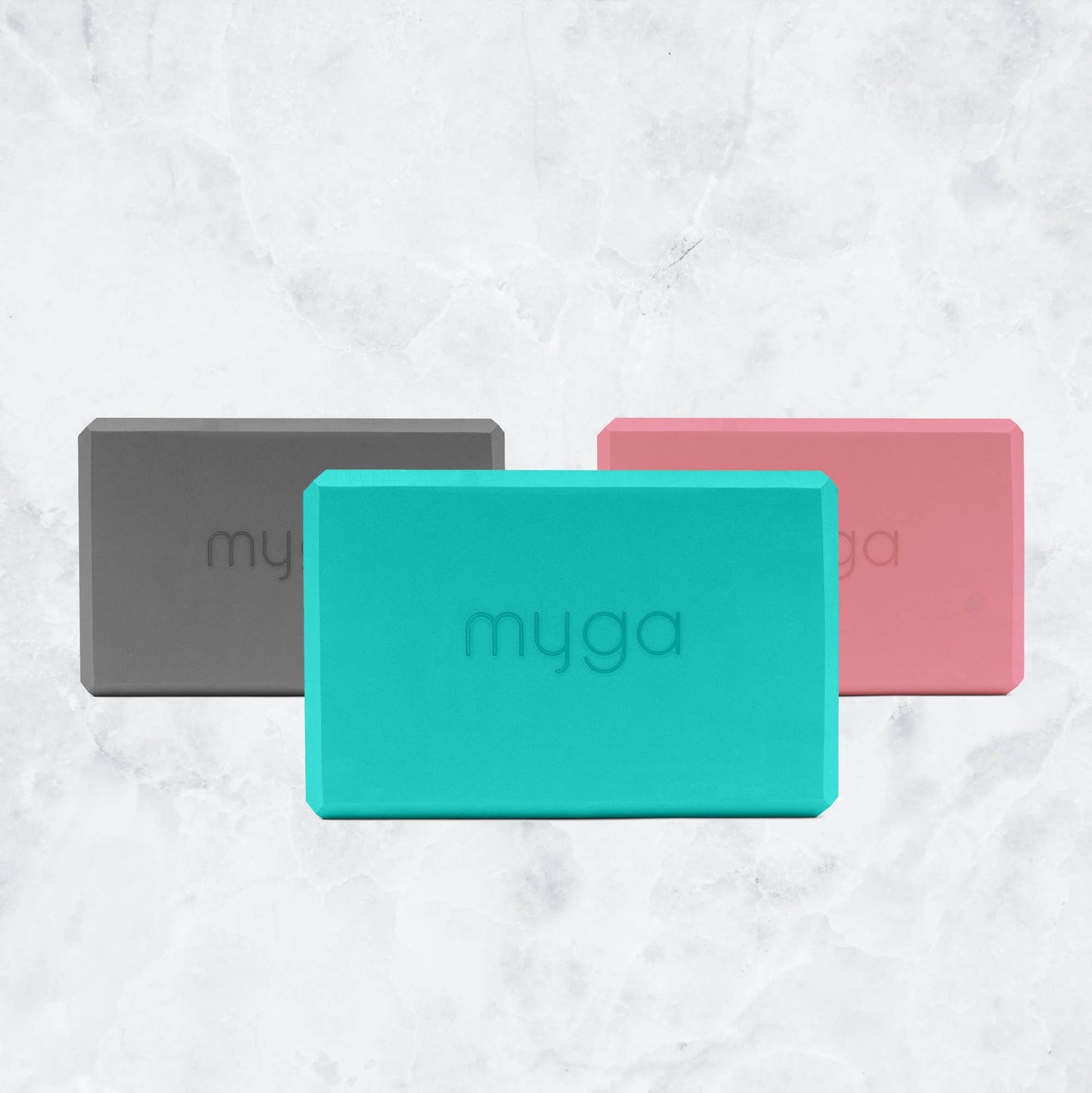 Three Myga foam yoga blocks are displayed together in a fan pattern on a marble background, showcasing the available colors: grey, teal/mint green, and pink. Each block has the Myga logo embossed on the front.