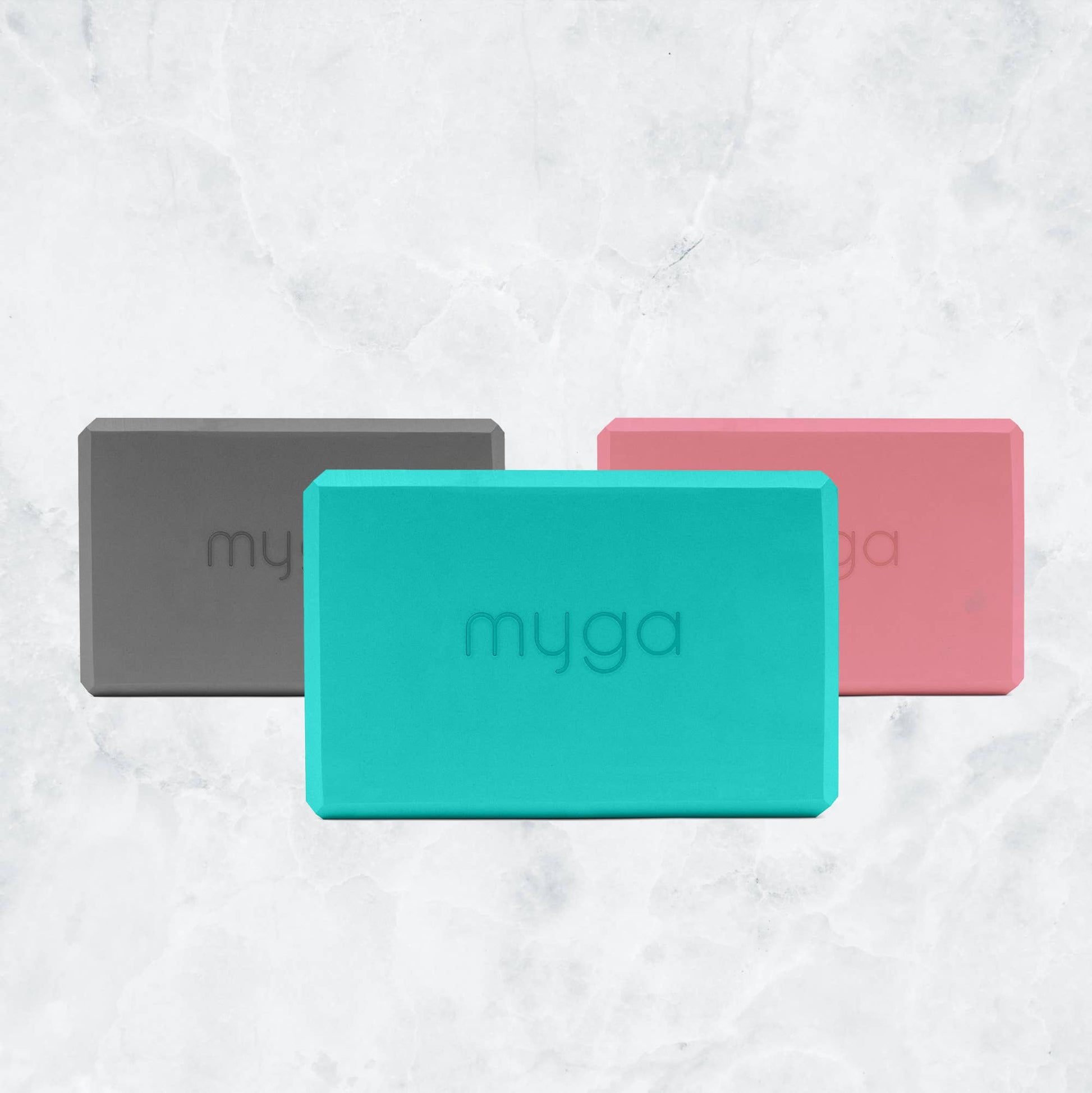 Three Myga foam yoga blocks are displayed together in a fan pattern on a marble background, showcasing the available colors: grey, teal/mint green, and pink. Each block has the Myga logo embossed on the front.