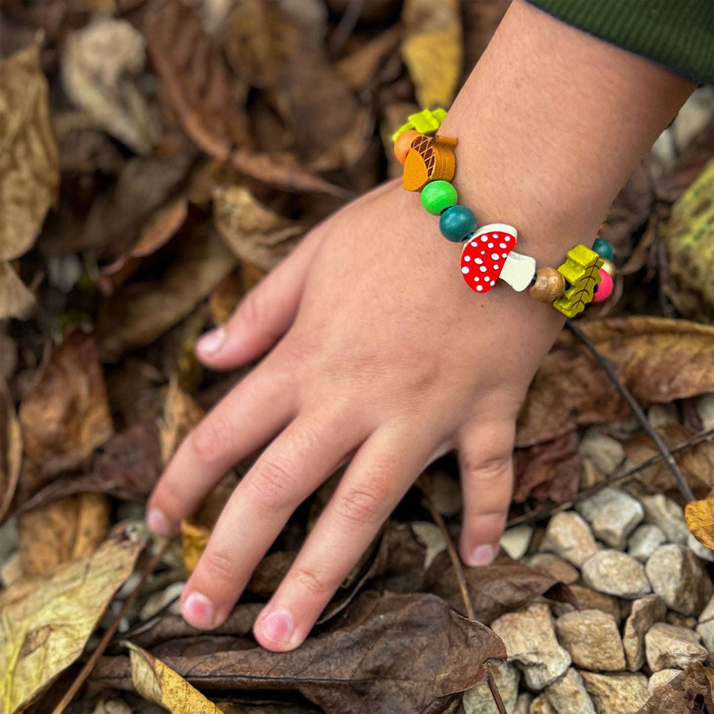 A child's hand rests on a ground covered in dry, brown autumn leaves, wearing a finished 'Forest Bracelet Kit' creation. The bracelet features colorful wooden beads, a large red and white mushroom bead, and green and yellow maple leaf charms.