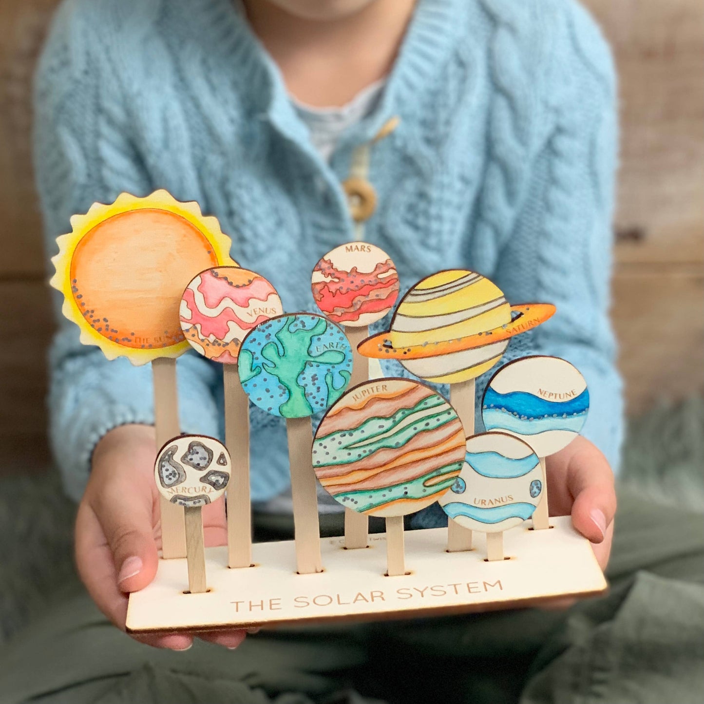 Child holding a finished, hand-painted wooden Solar System educational toy. The display includes the Sun and all eight planets (Mercury, Venus, Earth, Mars, Jupiter, Saturn, Uranus, Neptune) on individual wooden sticks, showcasing a fun, DIY space craft kit.