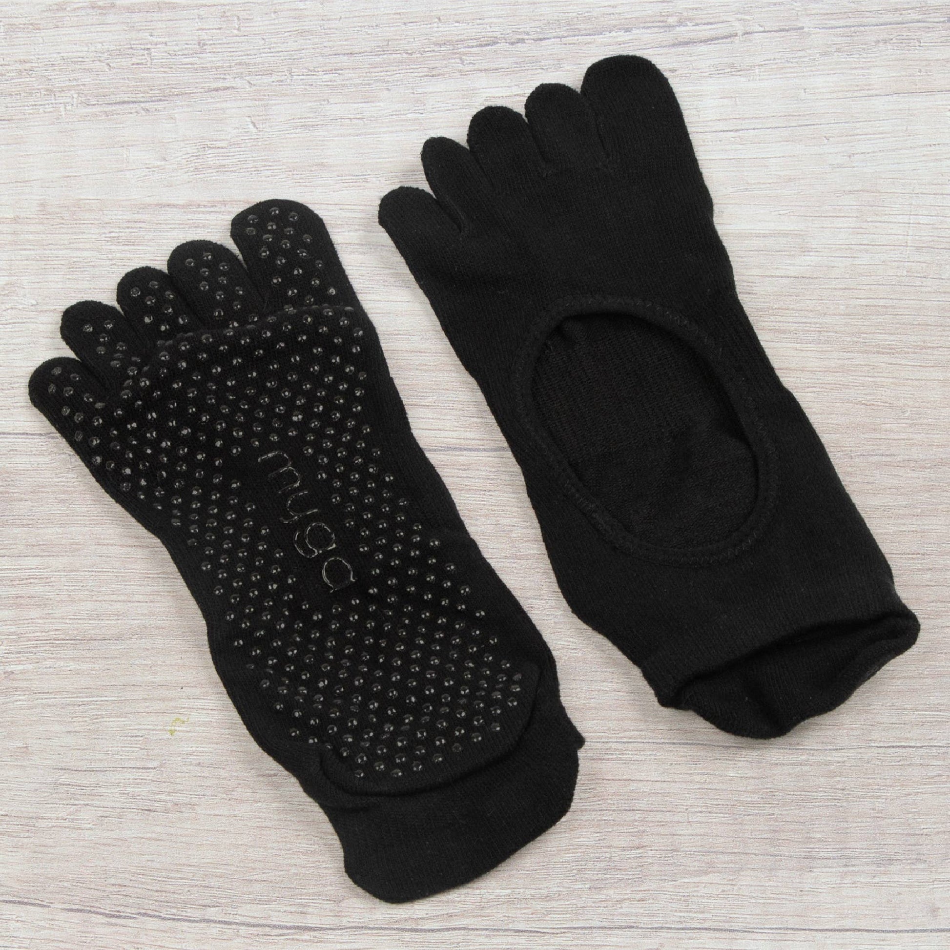 A pair of black full-toe grip socks by Myga, laid out on a light wooden surface. One sock is flipped to clearly show the non-slip dots on the sole, while the other shows the smooth top and the large arch cutout.