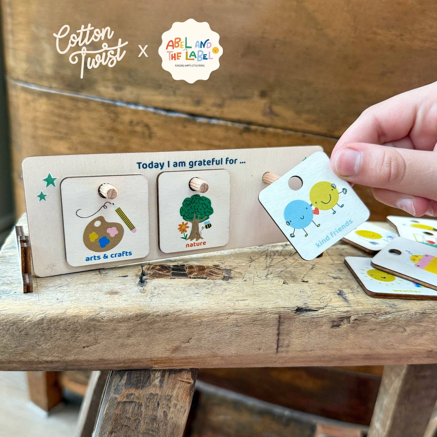 A hand places the Good Friends token onto the wooden "Today I am grateful for" chart, a collaboration between Cotton Twist and ABL. The display currently shows the Arts & Crafts and Nature tokens. The gratitude chart sits on a rustic wooden table, showcasing the product in a home setting.