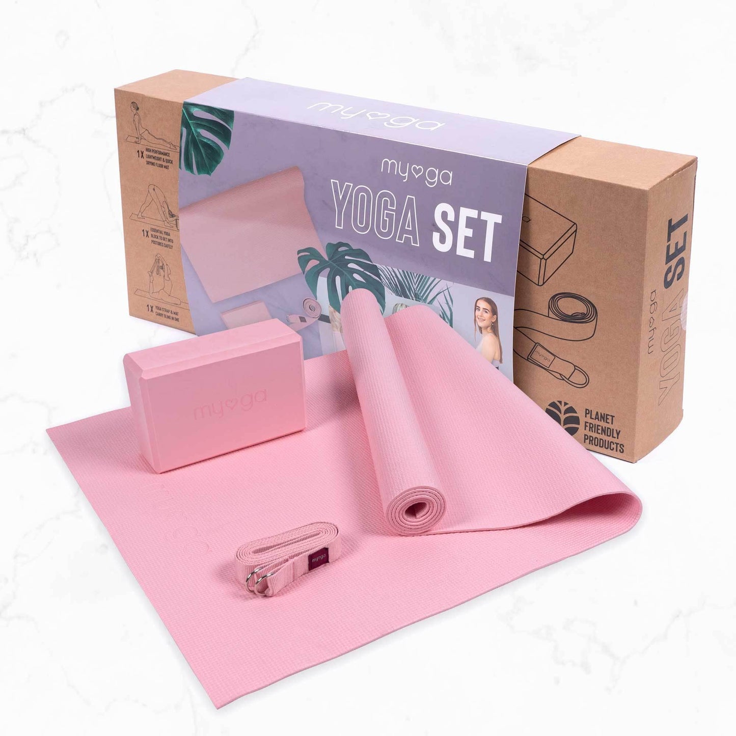 The Myga 3-piece Yoga Set in light pink is displayed on a marble-style floor in front of its cardboard retail box. The set includes a rolled yoga mat, a foam yoga block, and a matching yoga strap, highlighting a complete kit for yoga or fitness practice.