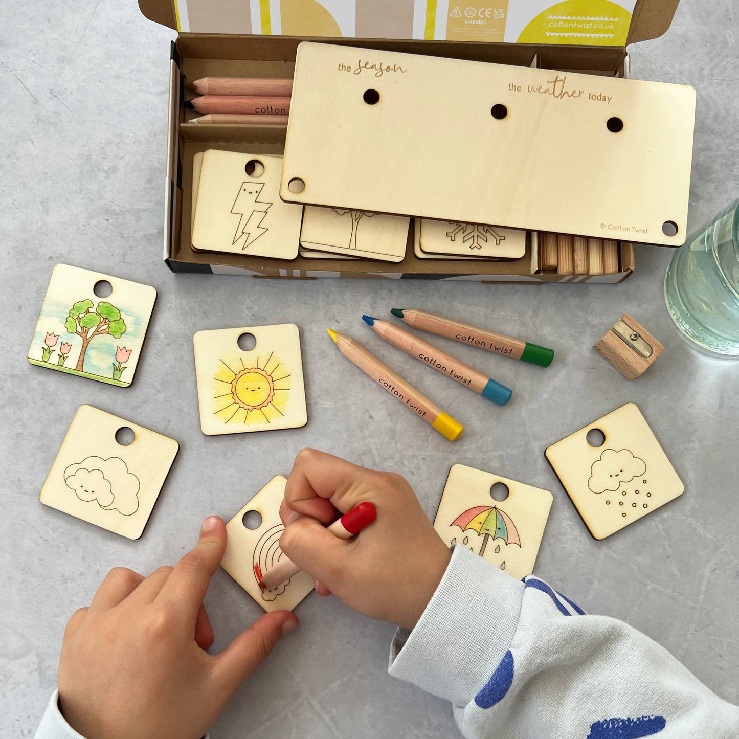 A child's hands color a wooden rainbow tag with a red pencil from the DIY weather and season chart craft kit. The open kit box contains a wooden display stand, colored pencils, and several other partially colored wooden tokens, including a sun, clouds, and a tree, spread out on a gray surface