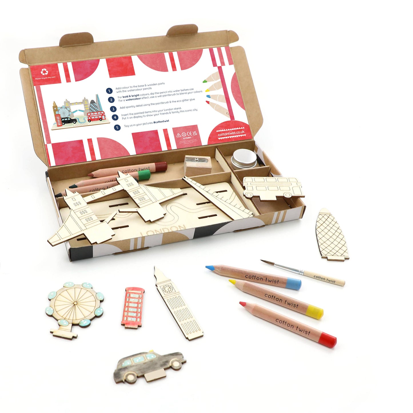 The open box for the DIY London Landmarks craft kit, showing all components: wooden pieces for Tower Bridge, Big Ben, London Eye, bus, and taxi; colored pencils; a sharpener; and the instructions. This is a complete children's build and color model set.