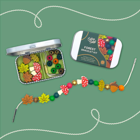 'Forest Bracelet Kit' by Cotton Twist, shown on a green background. The open tin contains green, yellow, and red wooden beads, thread, and unique beads shaped like mushrooms and leaves. A finished bracelet with mushroom and autumn leaf beads is displayed in the foreground, ready for threading