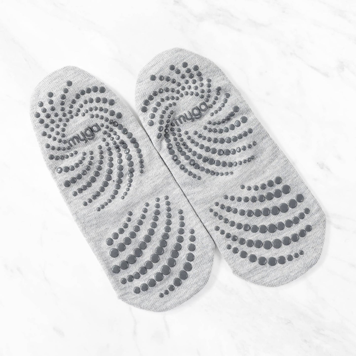 Overhead view of a pair of grey non-slip yoga or barre socks laid out, showing the entire sole. The soles feature a distinct swirling pattern of dark grey grip dots to ensure maximum traction and stability during floor work.