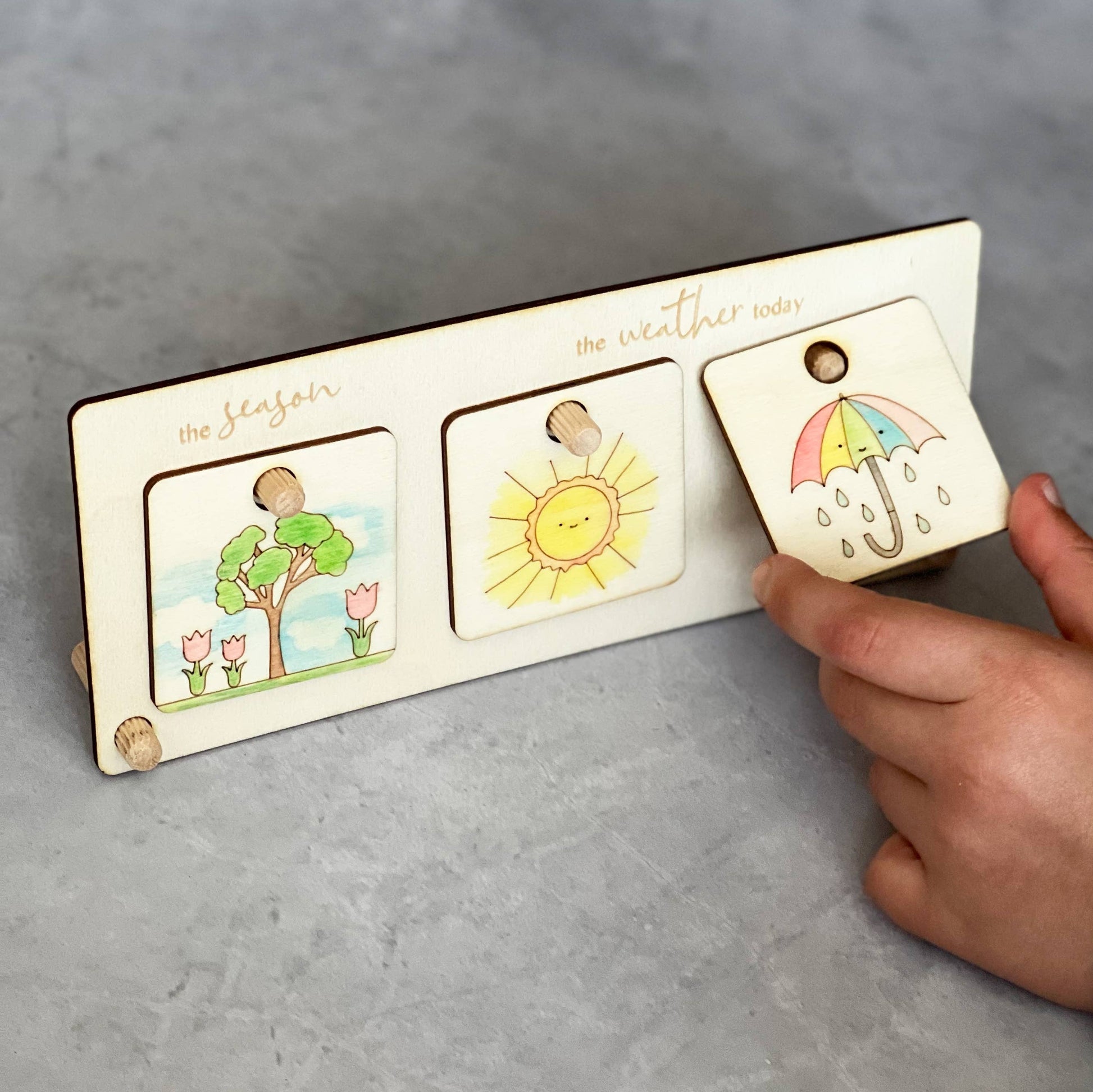 A hand is placing a hand-colored wooden umbrella token onto the DIY weather chart stand. The chart currently displays two other completed tokens: a flowering tree for 'the season' and a smiling sun for 'the weather today.' This shows the wooden educational chart in use.
