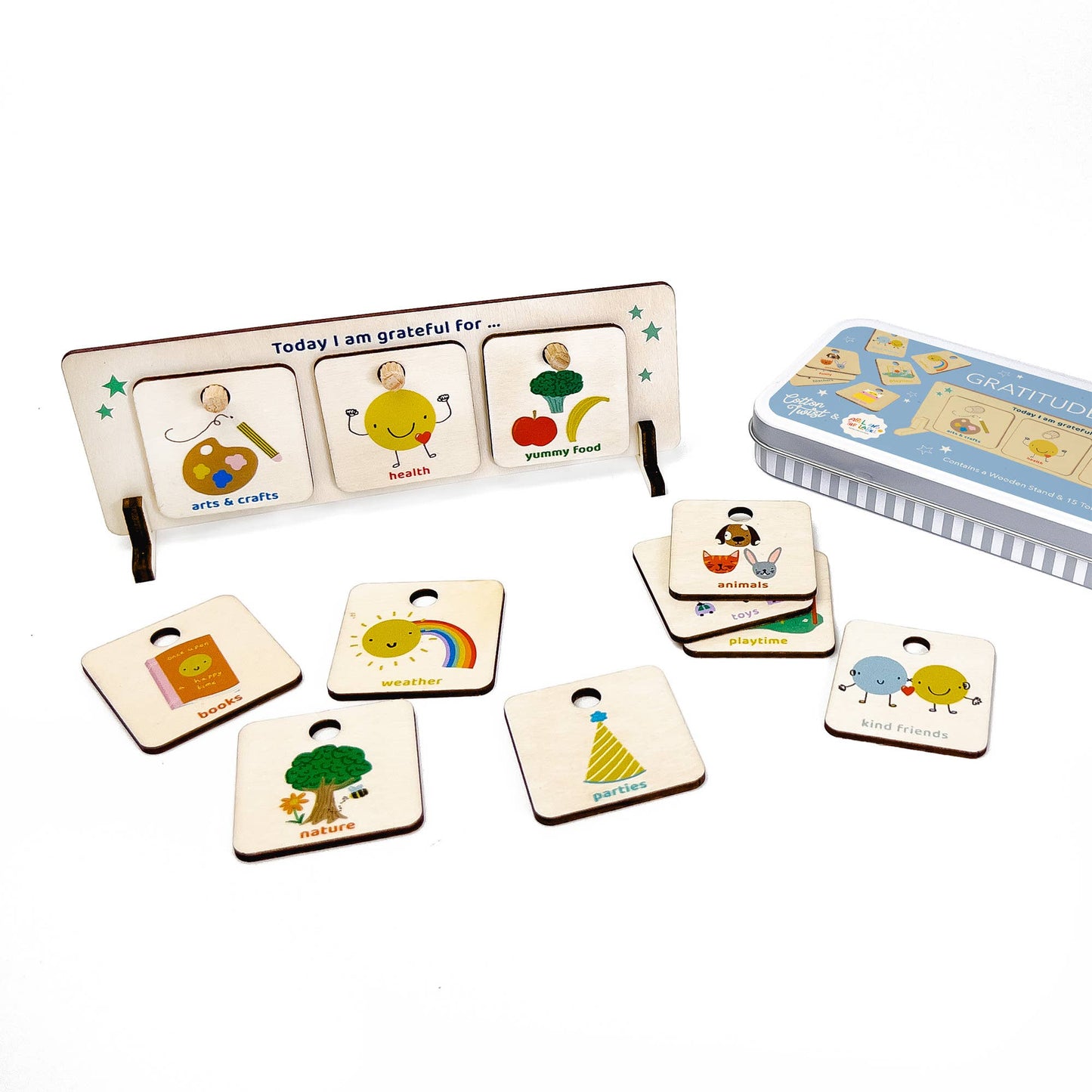 The complete wooden Gratitude Chart set is displayed next to its blue storage tin. The stand shows three tokens: Arts & Crafts, Health, and Yummy Food. Scattered below are additional tokens, including Family, Nature, Sun/Rainbow, Parties, and Reading/Books, showcasing the full set of gratitude tokens.