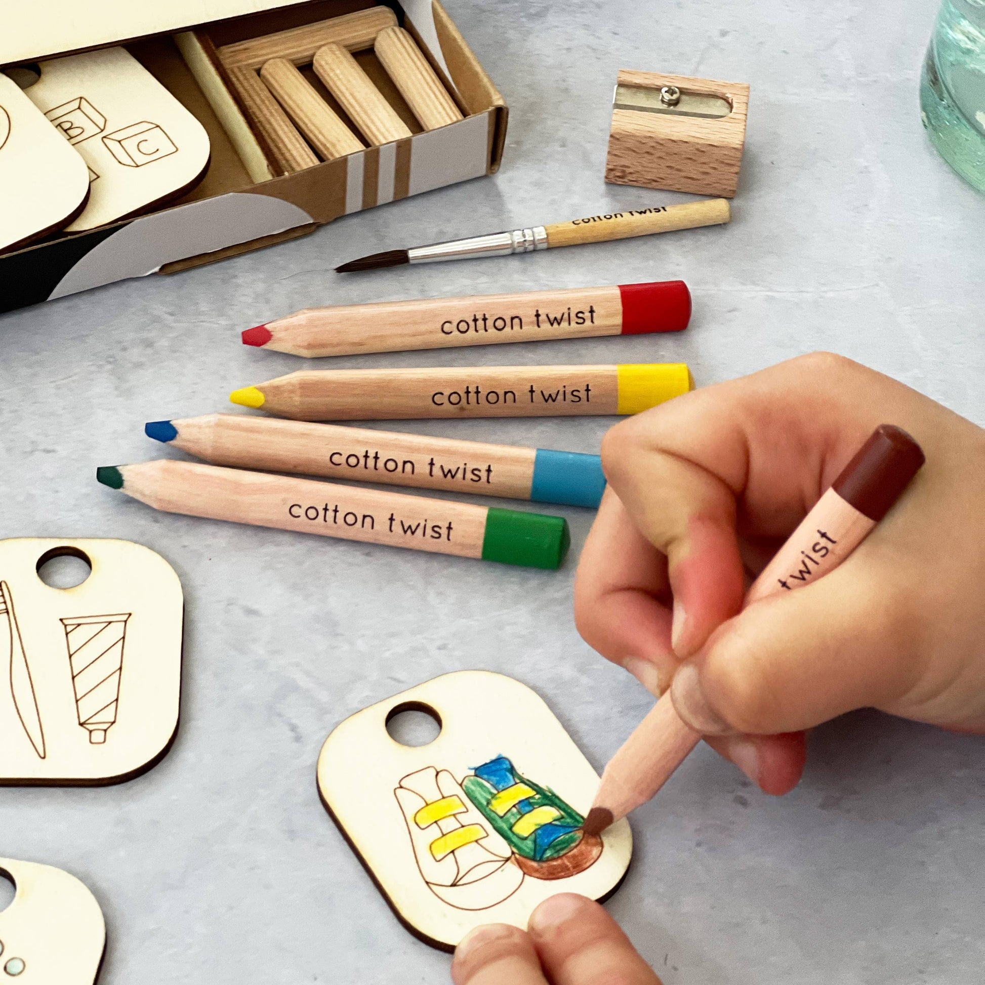 Close-up of a child's hands coloring the wooden getting dressed token (shoes and socks) with a red Cotton Twist branded pencil from the Daily Task Chart kit. Uncolored chore tokens, including brushing teeth and a tube of toothpaste, are laid out, showing the hands-on activity and kit components.