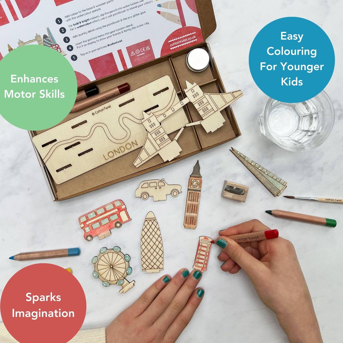 Marketing graphic showing a child coloring a Tower Bridge piece from the DIY London Landmarks craft kit, highlighting the developmental benefits: Enhances Motor Skills, Easy Colouring For Younger Kids, and Sparks Imagination. The open kit box, wooden stand, and other uncolored landmark pieces are visible in the background.