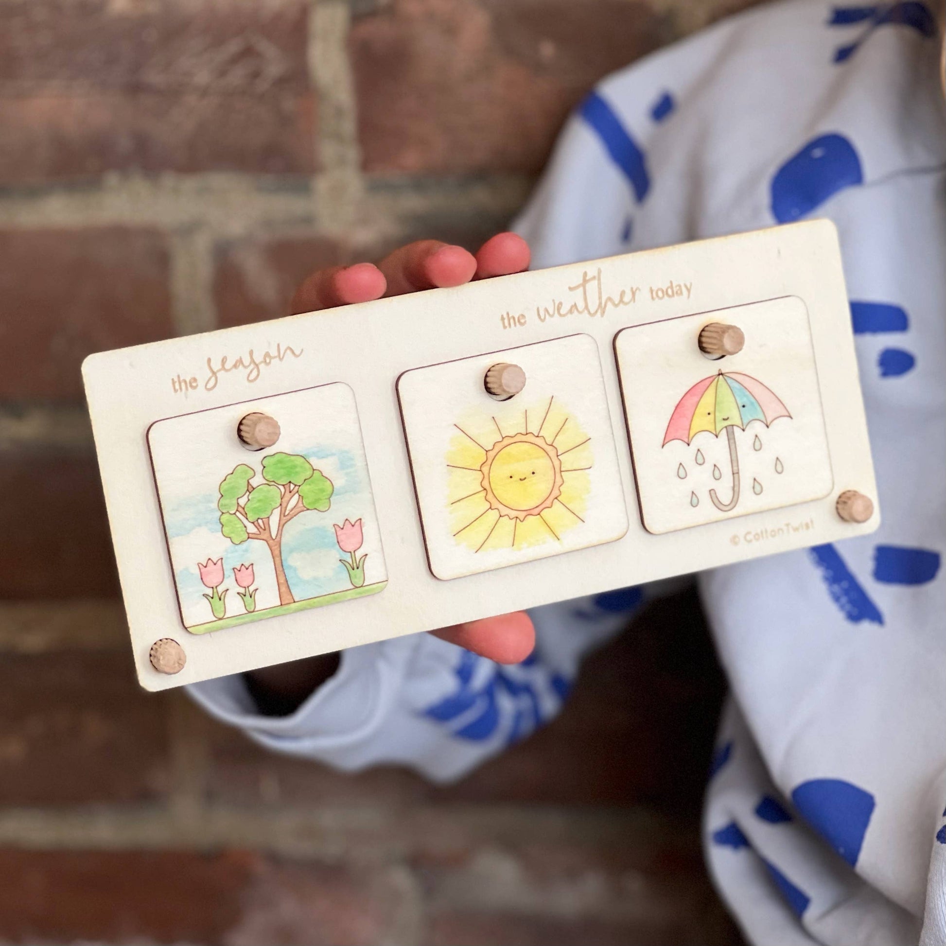 Wooden weather and season chart held by a child. The stand displays three hand-colored wooden tags: a tree with flowers representing 'the season,' a smiling sun for 'the weather today,' and a colorful umbrella and raindrops. This demonstrates the completed craft kit.