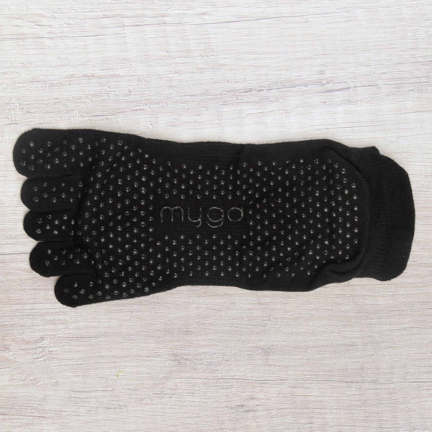 Overhead view of a single black full-toe grip sock by Myga. The sock is spread flat, clearly showcasing the dense pattern of non-slip grip dots on the sole for enhanced traction during yoga or Pilates practice.