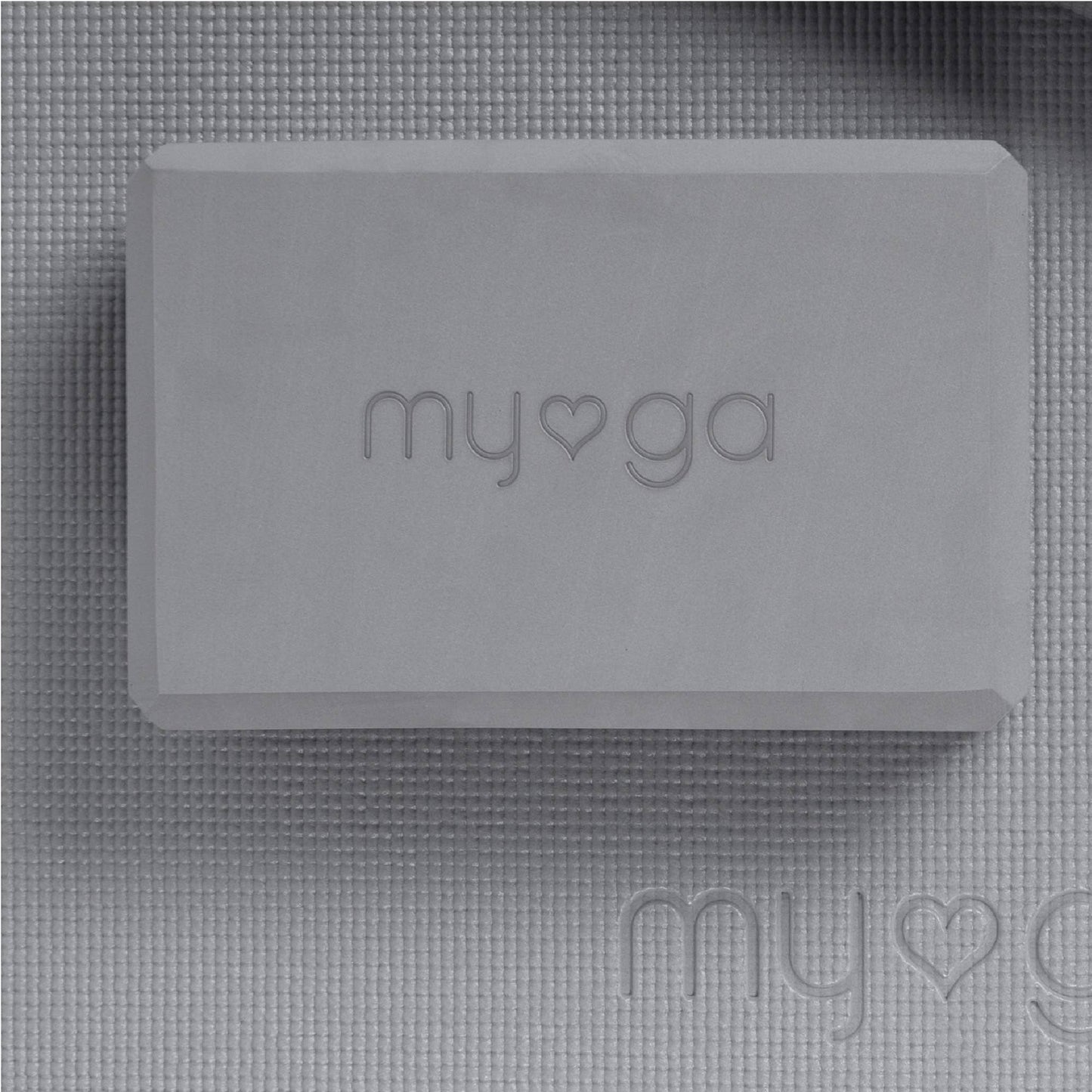 Extreme close-up overhead shot of a grey Myga foam yoga block. The block, embossed with the Myga logo, rests on a matching grey yoga mat, showcasing the texture of the accessories.