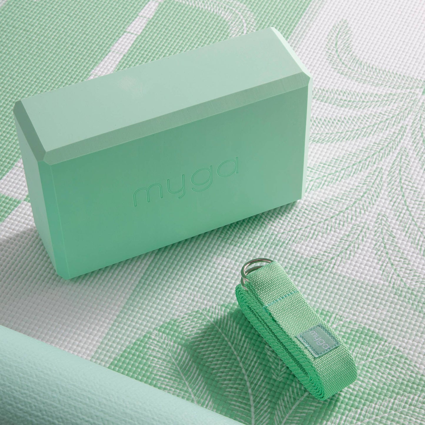 Close-up of a sage palm Myga foam yoga block and a folded yoga strap with a metal ring, resting on a patterned green and white yoga mat. This shot highlights the accessories for a fitness practice.