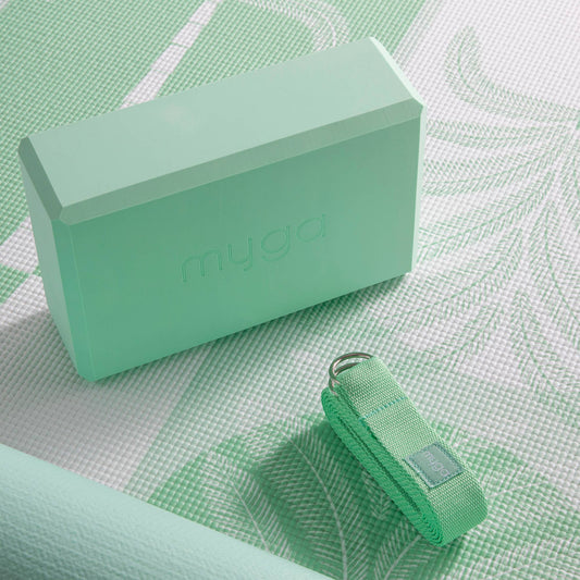 Close-up of a sage palm Myga foam yoga block and a folded yoga strap with a metal ring, resting on a patterned green and white yoga mat. This shot highlights the accessories for a fitness practice.