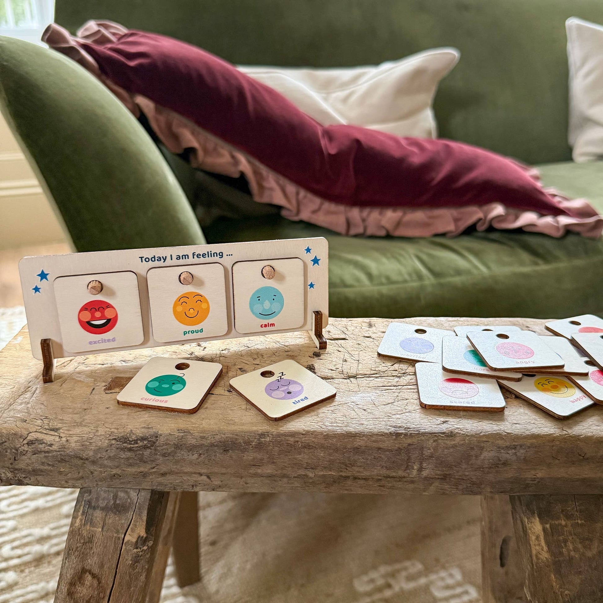 Wooden feelings chart set up on a rustic wooden table. The display shows 'excited,' 'proud,' and 'calm' tiles. A full collection of interchangeable emotion tiles is scattered to the side. The scene is set in a home, with a green velvet sofa in the background, showing the product is for everyday use.