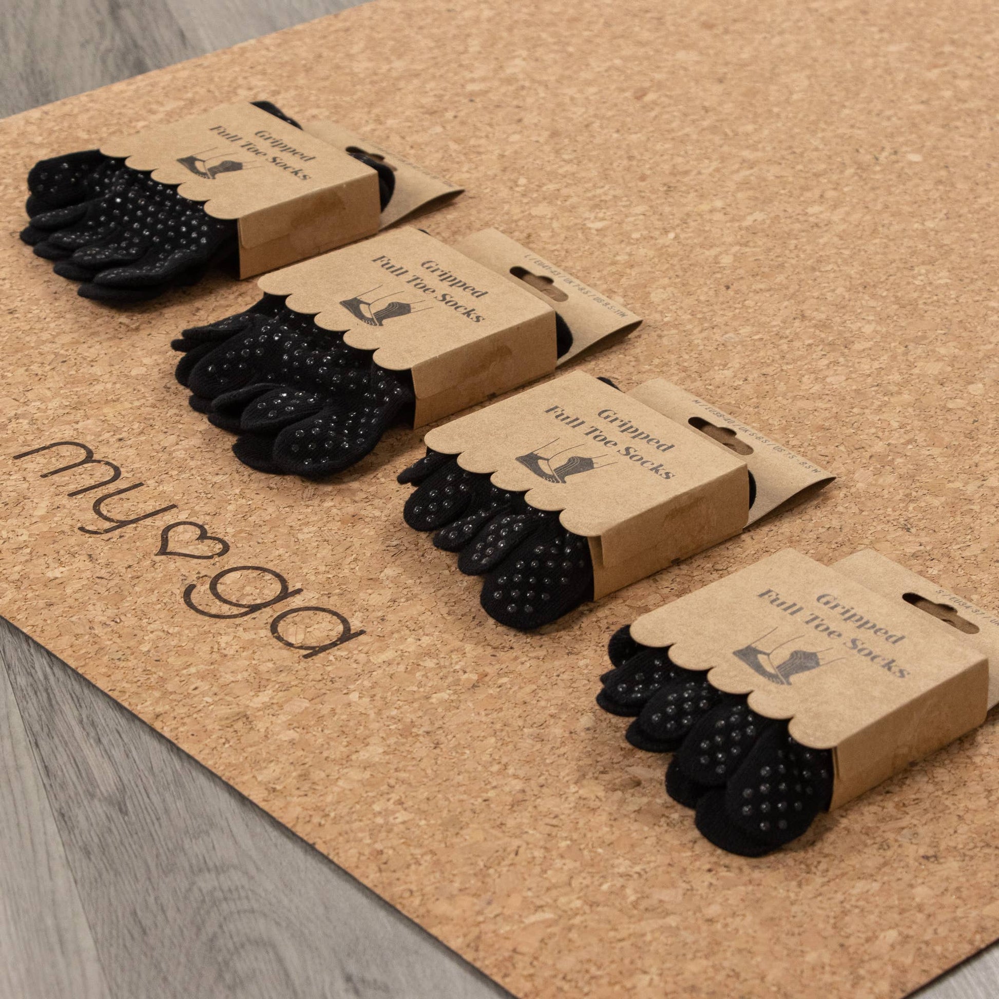 Four packages of Myga Gripped Full Toe Socks in black are displayed diagonally on a natural cork yoga mat. The brown cardboard packaging clearly shows the socks and the non-slip grip dots, emphasizing the product's function for yoga or Pilates.