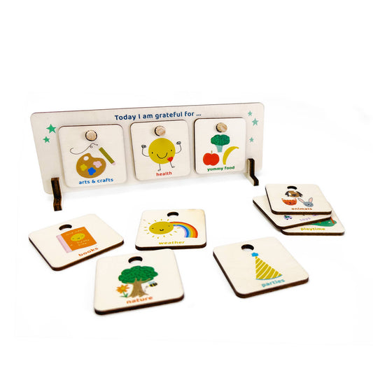 The complete wooden Gratitude Chart set, titled "Today I am grateful for," with the display stand showing three tokens: Arts & Crafts, Health, and Yummy Food. Several interchangeable tokens are spread around the base, including Family, Nature, Parties, and a Rainbow, showcasing the variety of gratitude options.