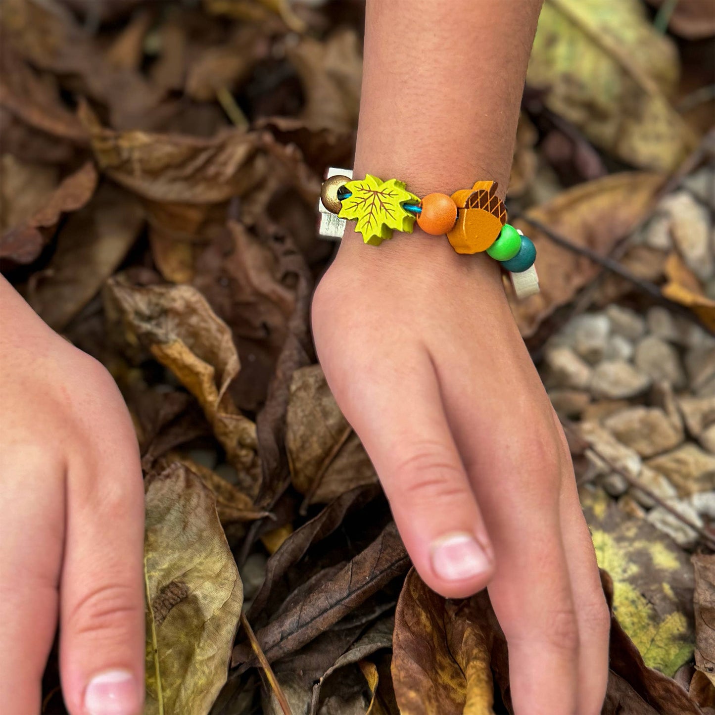 Close-up of a child's arm wearing a handmade bracelet from the Forest Bracelet Kit. The bracelet features vibrant wooden beads and forest-themed charms, including a bright green maple leaf and a brown acorn bead, against a backdrop of dark brown fallen leaves.