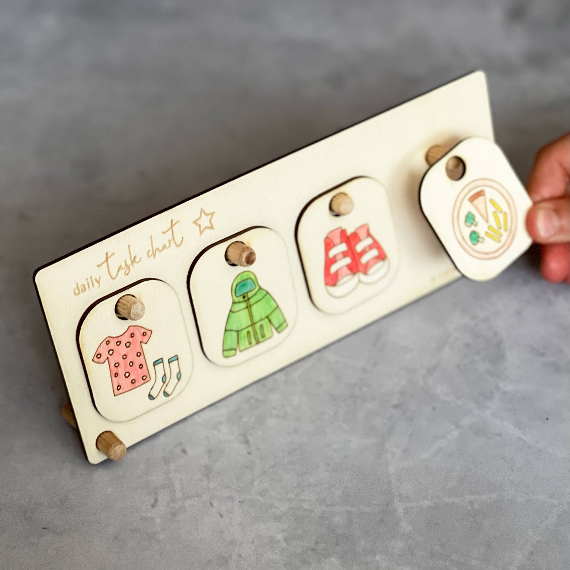 A hand is placing the hand-colored eating vegetables (or mealtime) token onto the wooden Daily Task Chart stand. The chart currently displays three other completed chore tokens: getting dressed (dress and socks), putting on a jacket, and putting on shoes, demonstrating the customizable routine board in use.