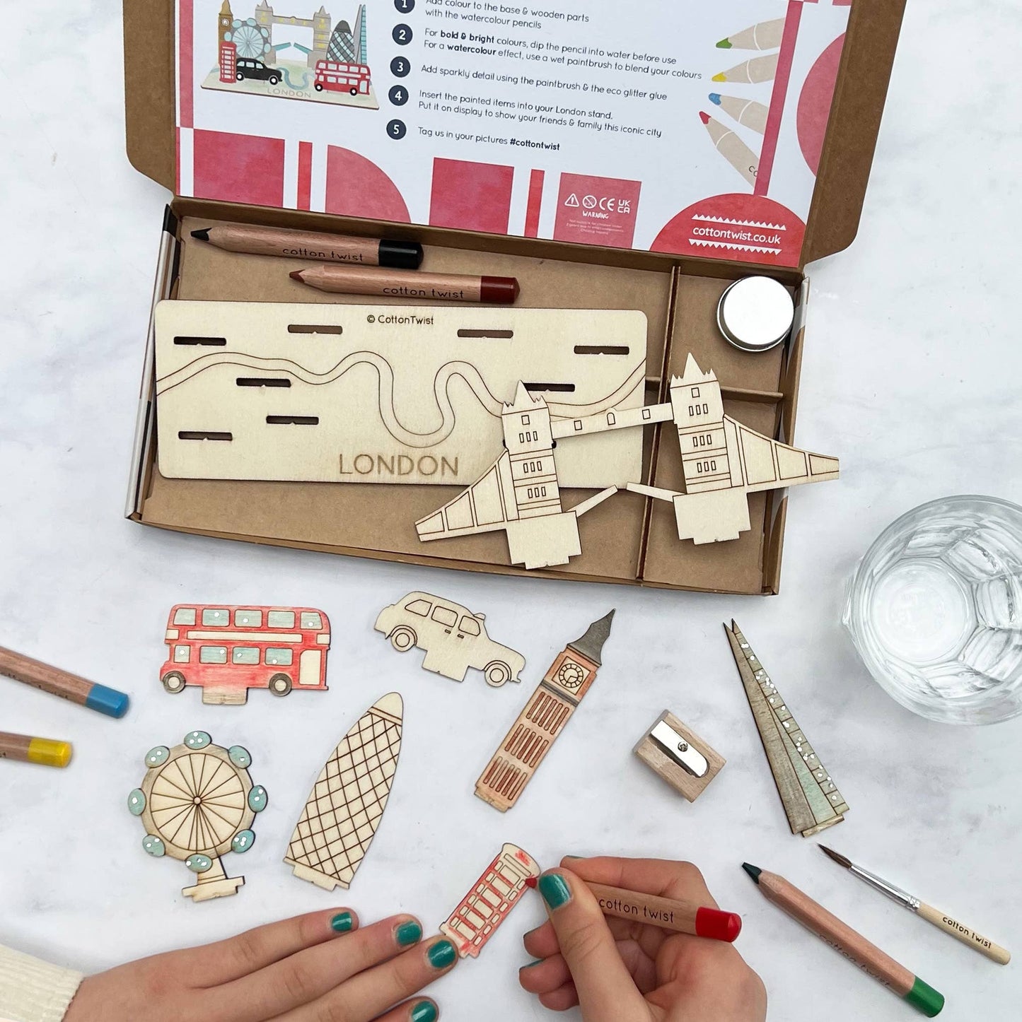 A child's hands are coloring a wooden red telephone booth piece from the DIY London Landmarks craft kit. The open box contains the unpainted 3D model base labeled 'LONDON.' Other pieces like Big Ben, the double-decker bus, and the London Eye are spread around, along with pencils and accessories, highlighting the hands-on activity.