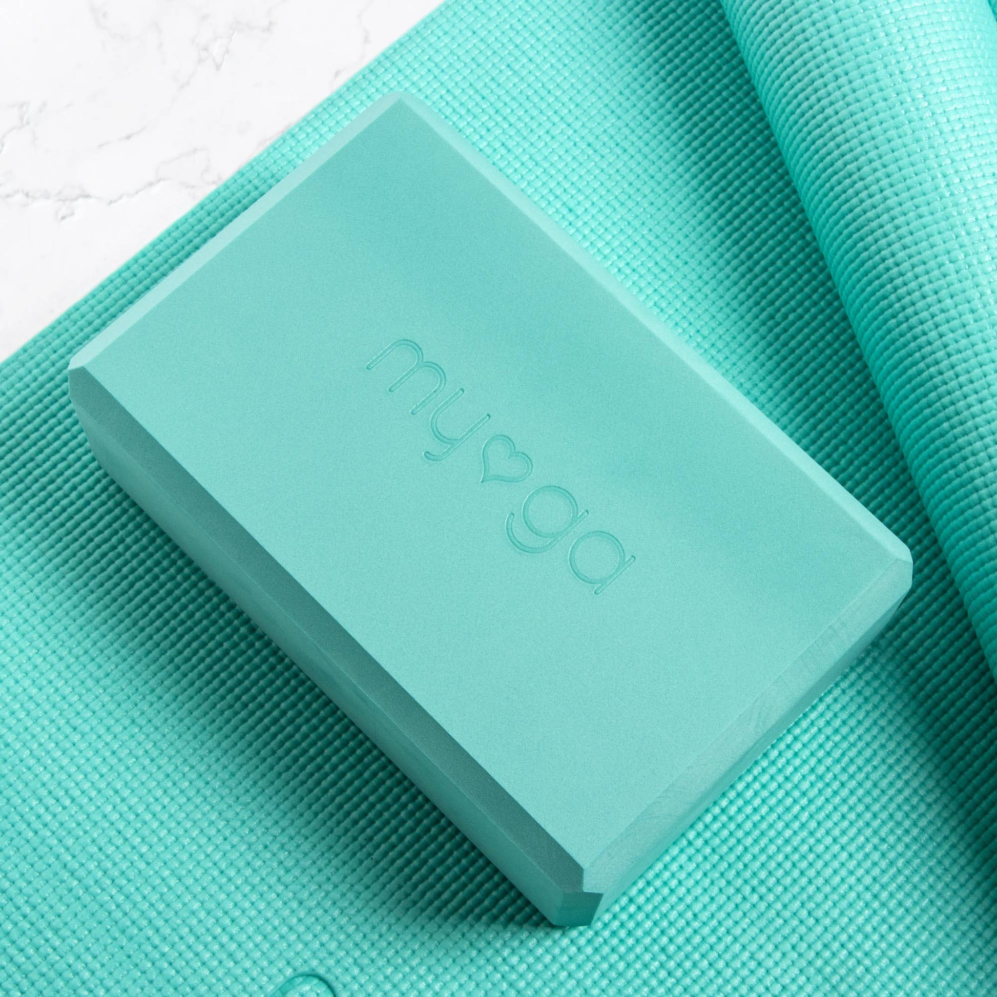 Extreme close-up of a teal/mint green Myga foam yoga block resting on a matching teal yoga mat. The block features the embossed Myga logo, showcasing the color and texture of the yoga accessory.