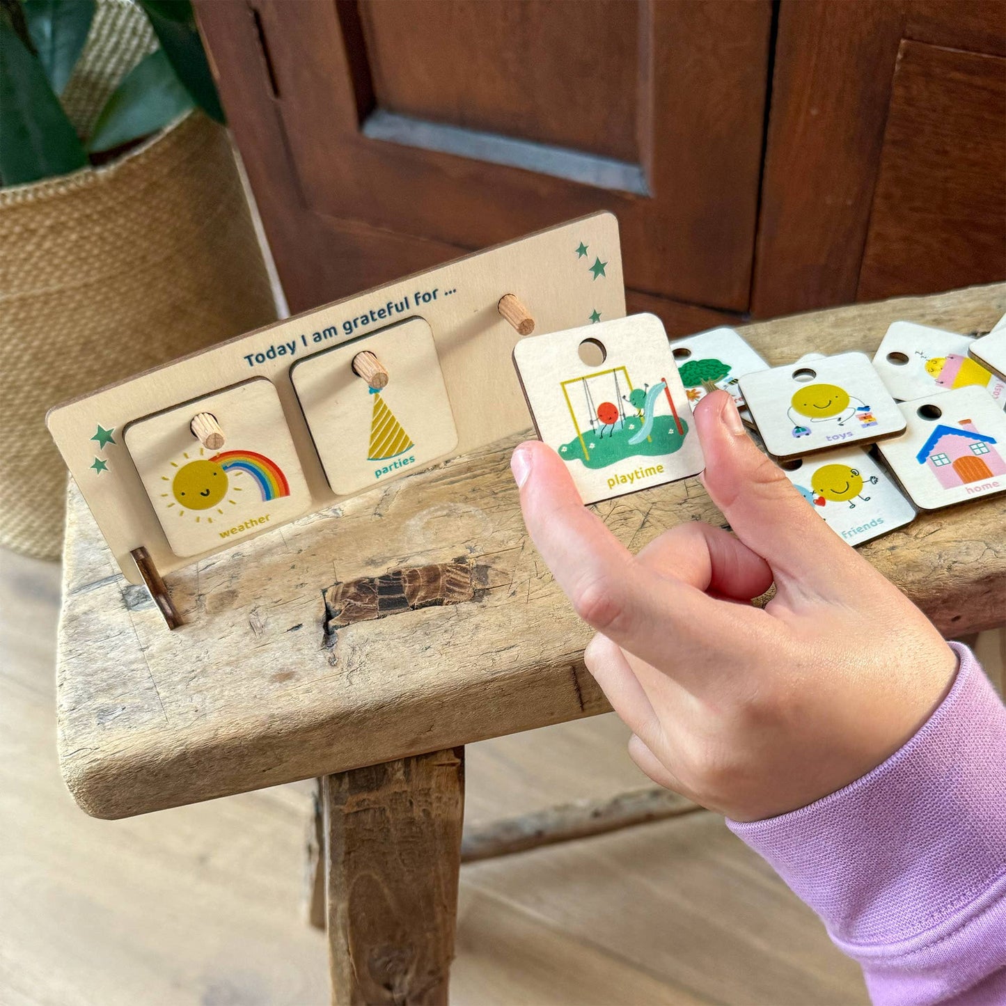 A child's finger points to the Playtime/Outdoors token being placed on the wooden "Today I am grateful for" chart. The stand currently displays the Sunshine/Rainbow and Parties tokens, highlighting the visual and interactive nature of the gratitude board. The chart rests on a rustic wooden bench.