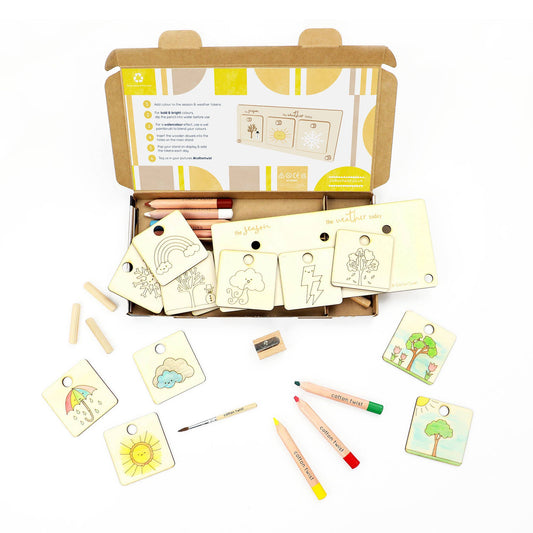 Open craft kit box containing wooden drawing or emotion tags, color pencils, a sharpener, and a small paintbrush. The blank wooden tags feature nature and weather-themed illustrations like a sun, rainbow, cloud, lightning bolt, and tree. The kit is designed for children to personalize the wooden pieces.
