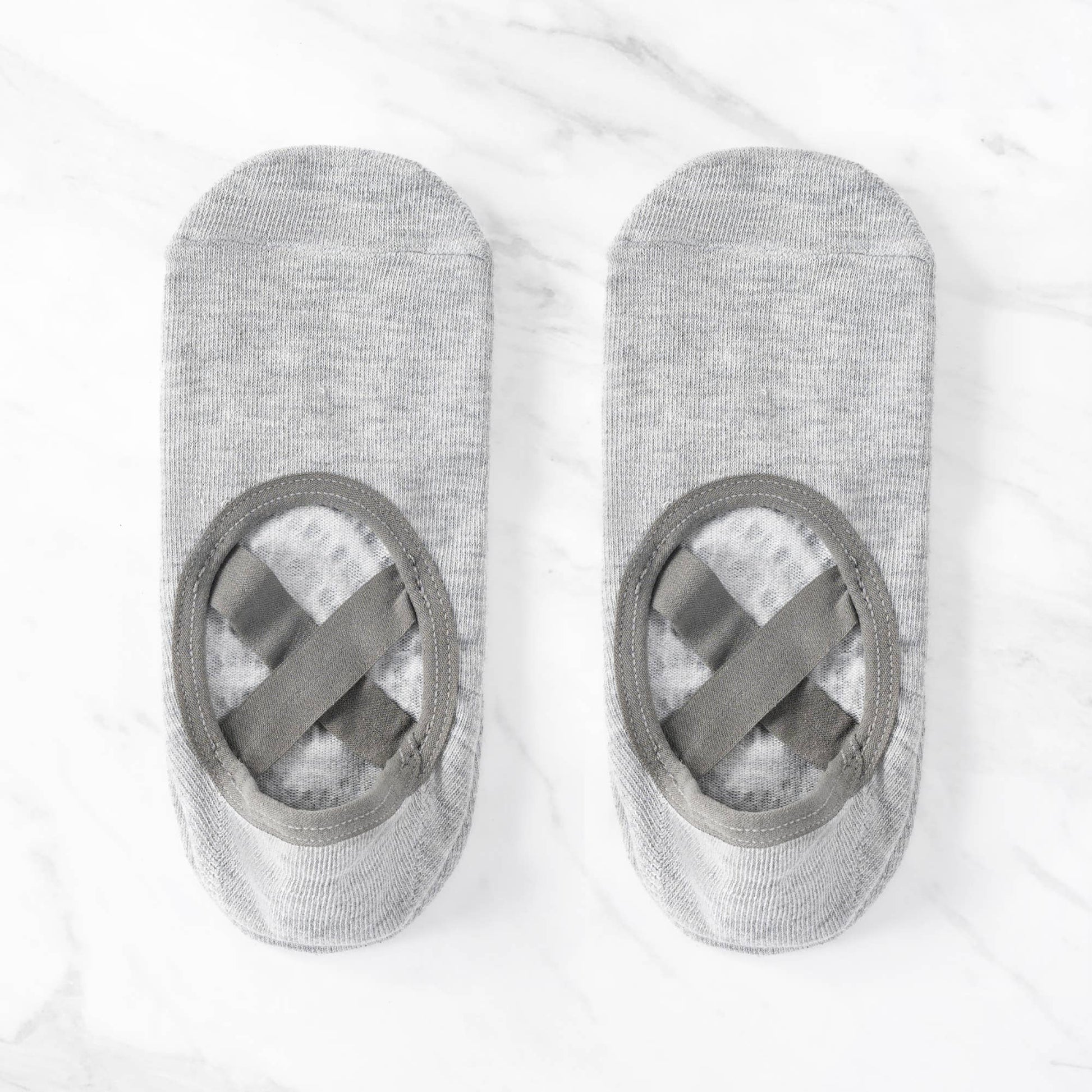 An overhead view of a pair of grey ballet-style non-slip grip socks. The socks feature a large open arch cutout with criss-cross elastic straps to keep them secure during yoga, Pilates, or barre practice.