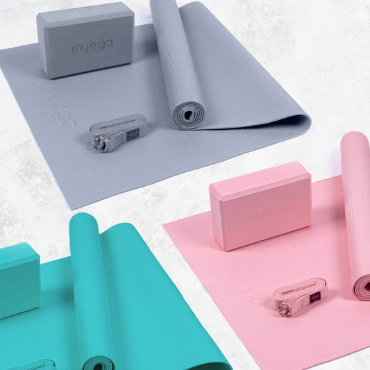 Overhead view of three complete Myga yoga sets in different colors: gray, pink, and teal. Each set includes a rolled yoga mat, a foam yoga block, and a yoga strap, showcasing a range of colorful yoga equipment.