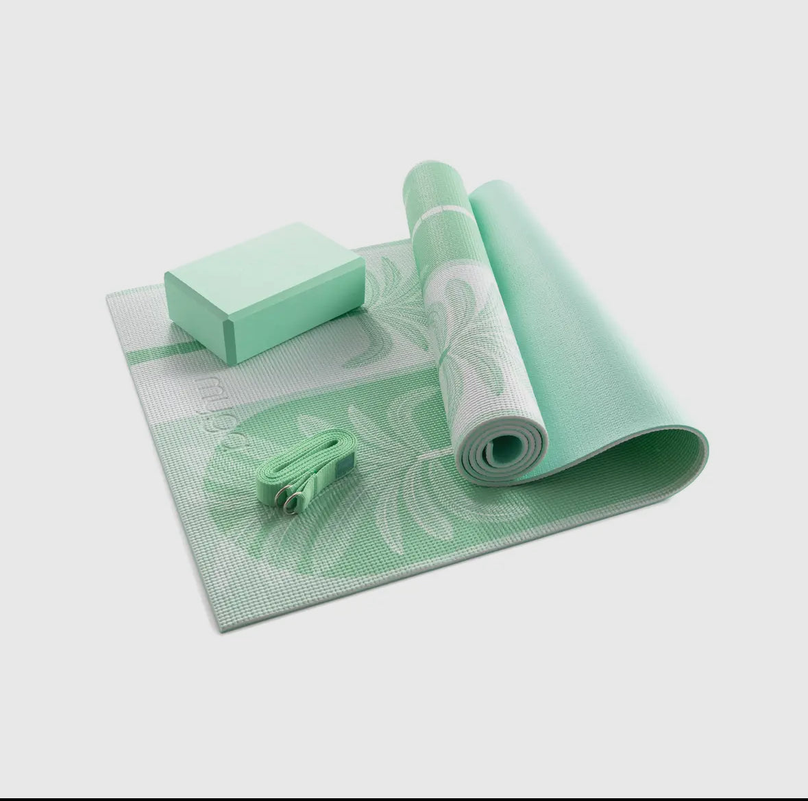 The Myga 3-piece Yoga Set in sage green is displayed, featuring a partially unrolled mat with a palm frond pattern, a foam yoga block, and a matching yoga strap. The set is showcased on a solid grey background.