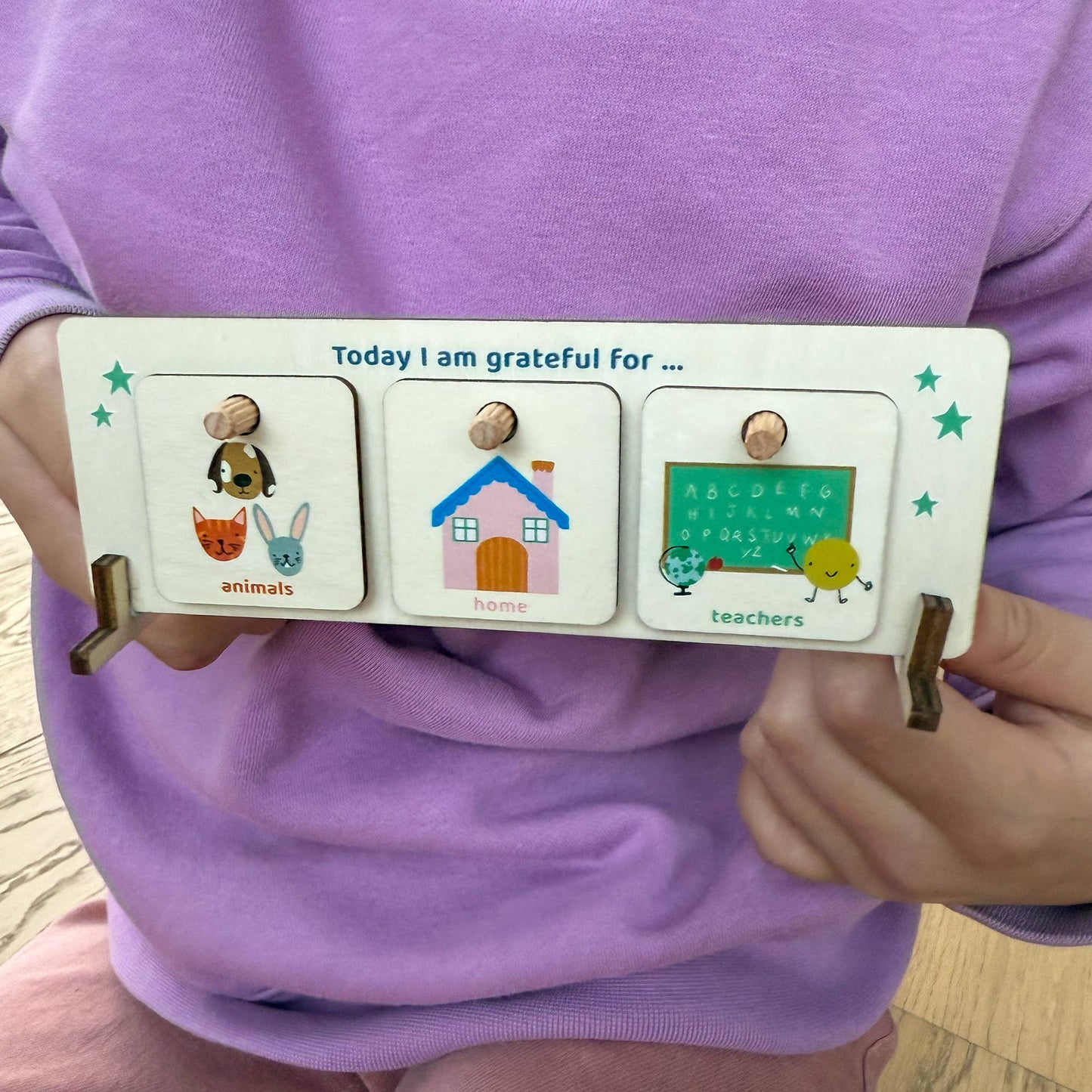 A child in a purple shirt holds the wooden Gratitude Chart, titled "Today I am grateful for." The display shows three tokens: Animals (mouse and rabbit), Home (a house), and Teachers (a pencil character), demonstrating the product's use as a tool for teaching thankfulness