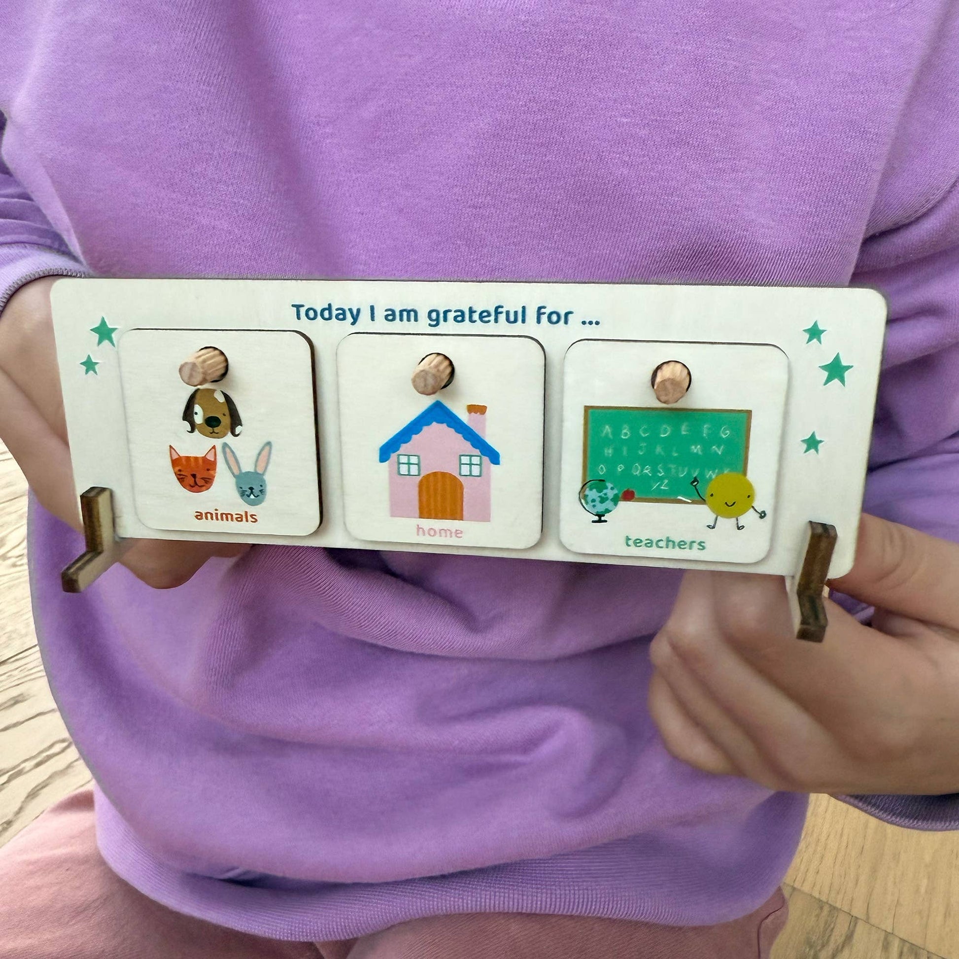A child in a purple shirt holds the wooden Gratitude Chart, titled "Today I am grateful for." The display shows three tokens: Animals (mouse and rabbit), Home (a house), and Teachers (a pencil character), demonstrating the product's use as a tool for teaching thankfulness