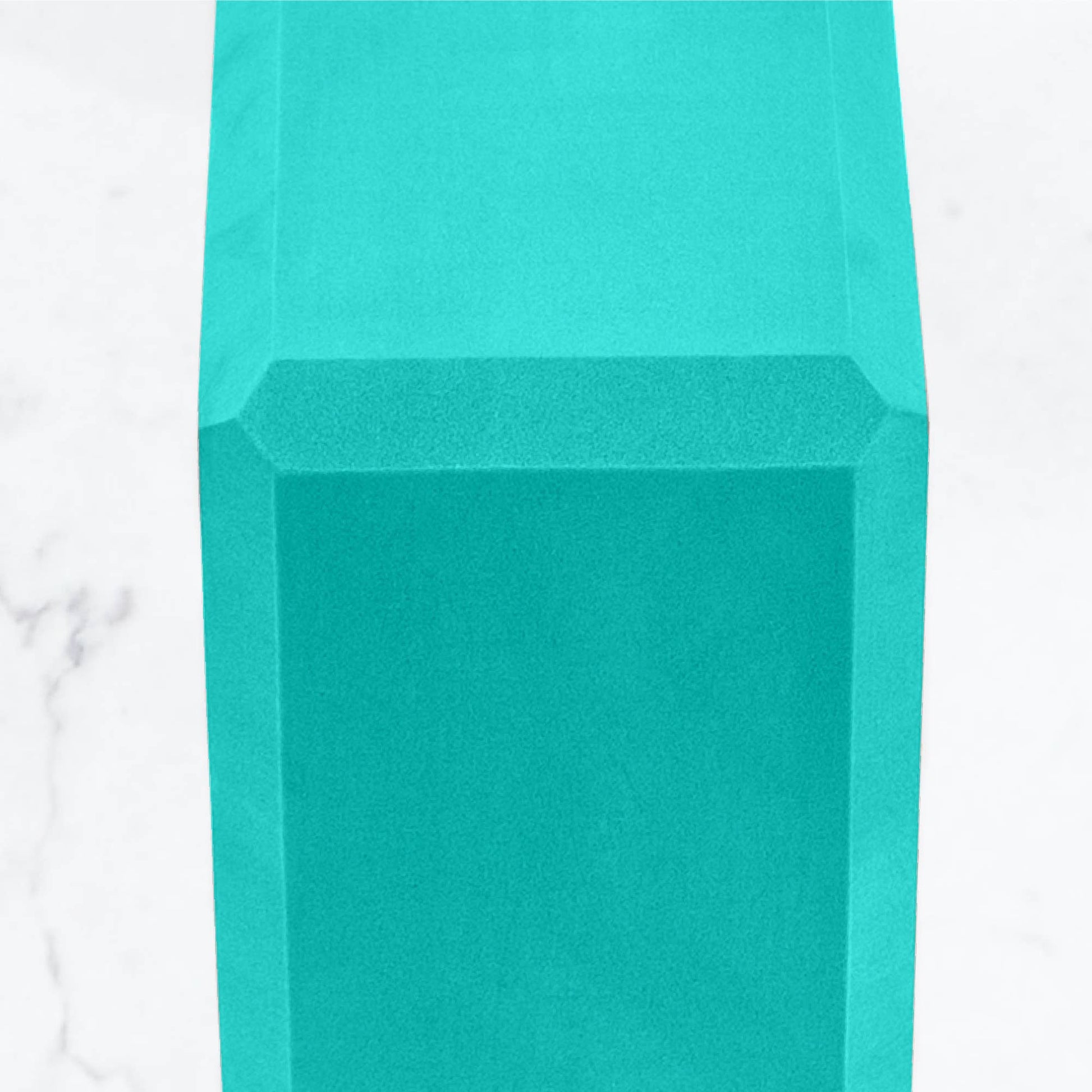 Extreme close-up on the side and top surface of a solid teal/mint green foam yoga block, highlighting the angled edges and smooth texture of the yoga prop on a marble background.