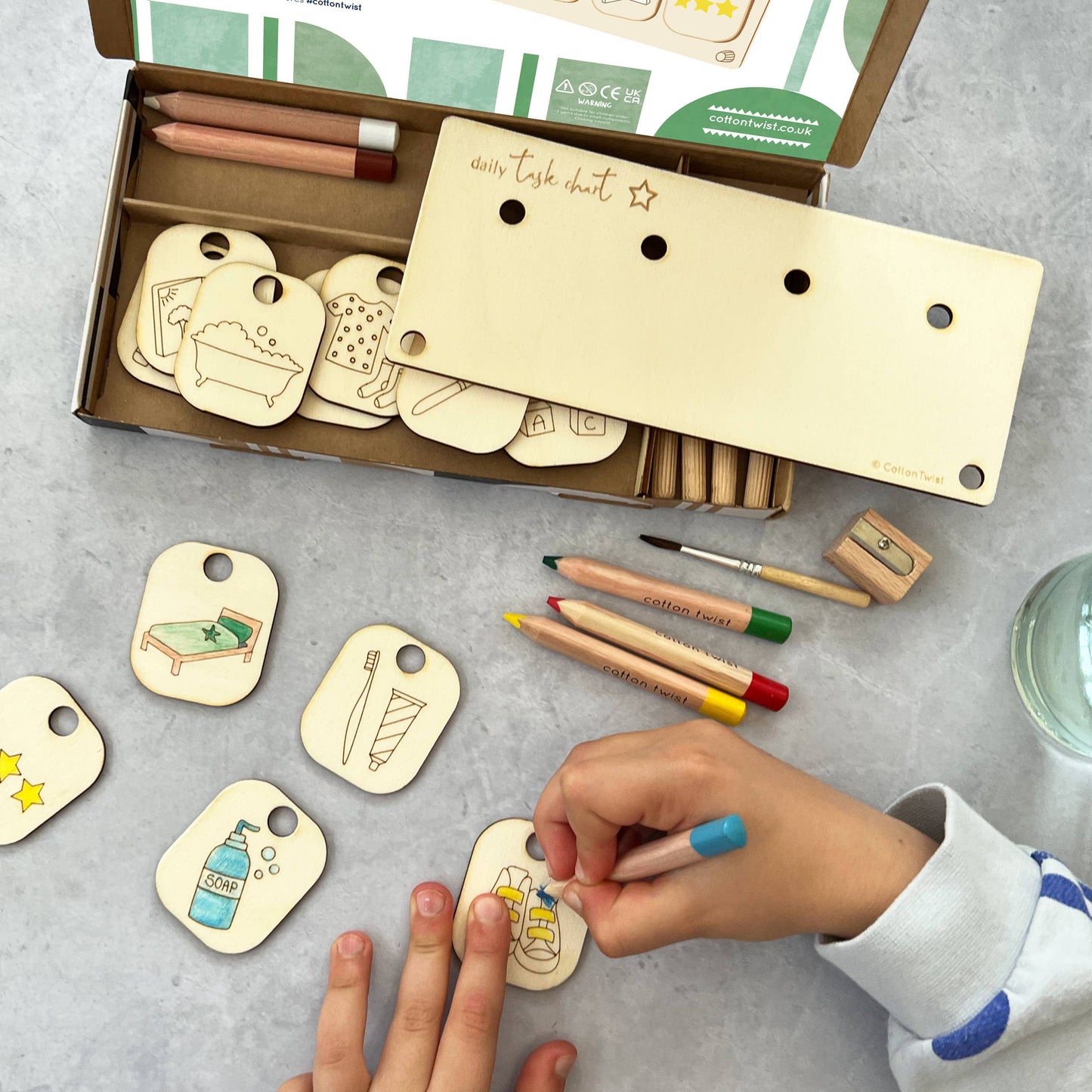 A child's hands are coloring a wooden chore token of a plate of food with a yellow pencil from the DIY Daily Task Chart craft kit. The open box, wooden stand, colored pencils, and several other blank and partially colored task tokens, including a made bed and a toothbrush, are spread on a gray table, illustrating the activity.