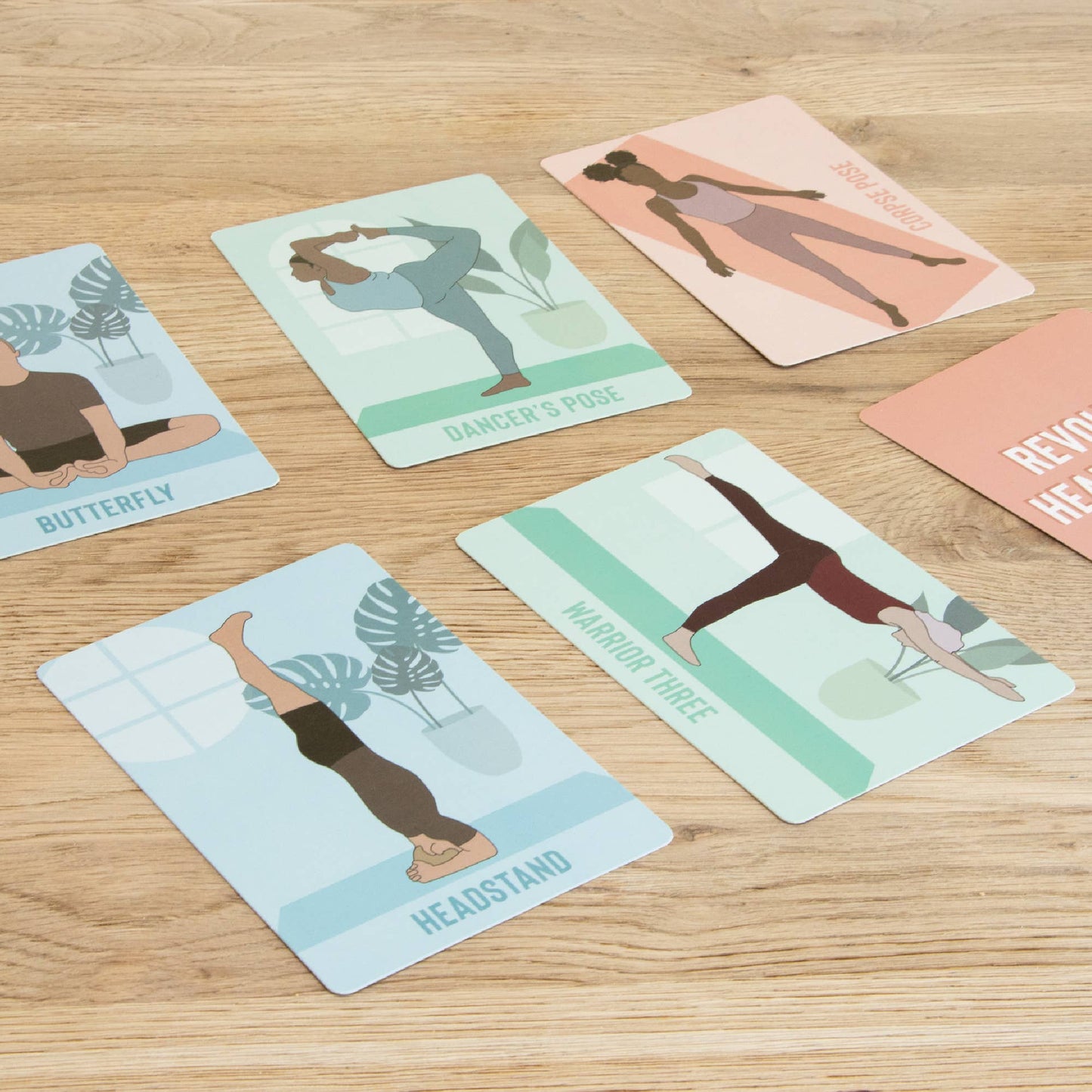 Multiple Myga Yoga Pose Cards are spread across a wooden table, featuring colorful illustrations and names of various poses, including Dancer's Pose, Warrior Three, Headstand, and Butterfly. The cards are instructional guides for yoga practice.