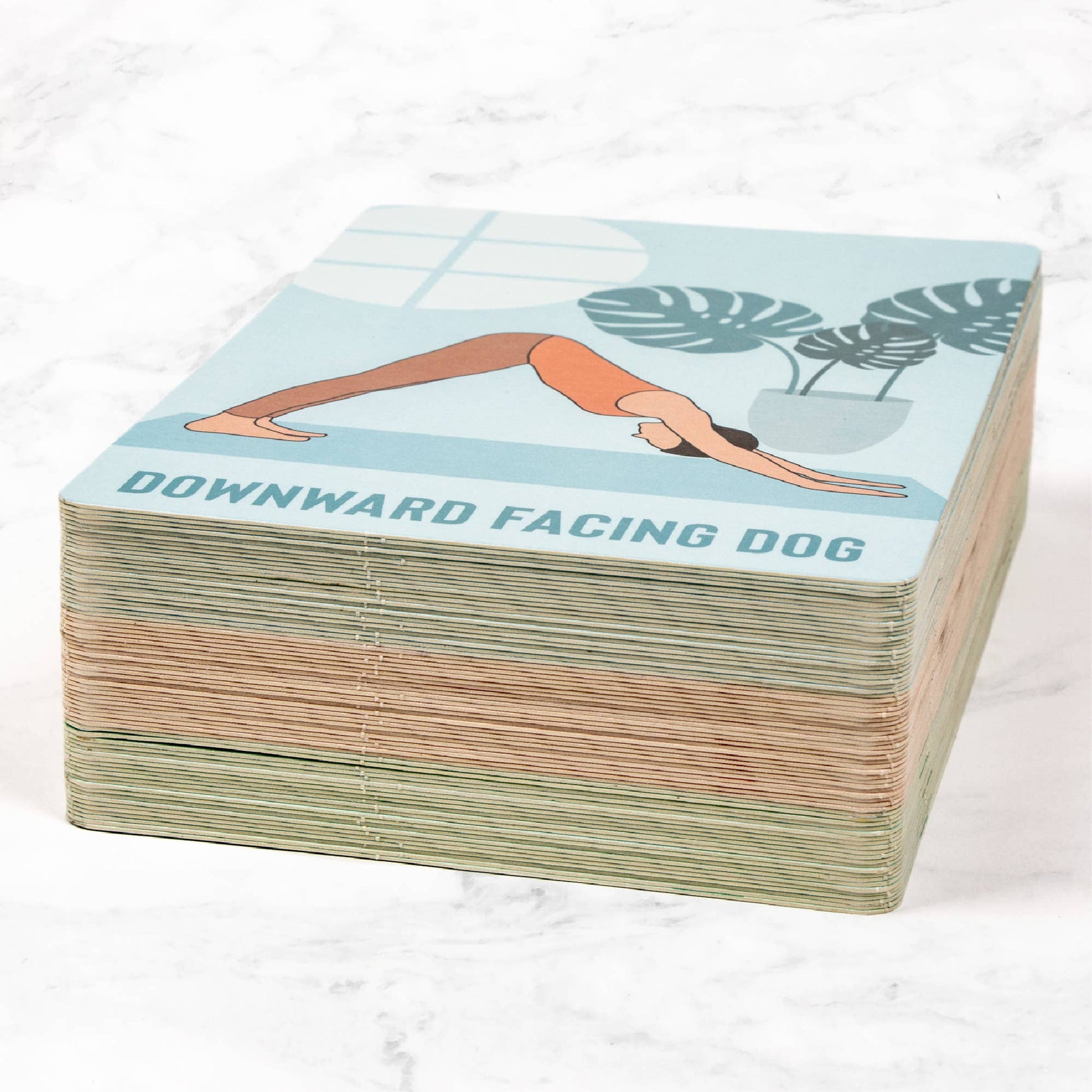 A full stack of the Myga Yoga Pose Cards with the top card showing the Downward Facing Dog pose illustration and name. The cards have colorful edges (blue, peach, and green), indicating the variety of poses in the instructional deck.