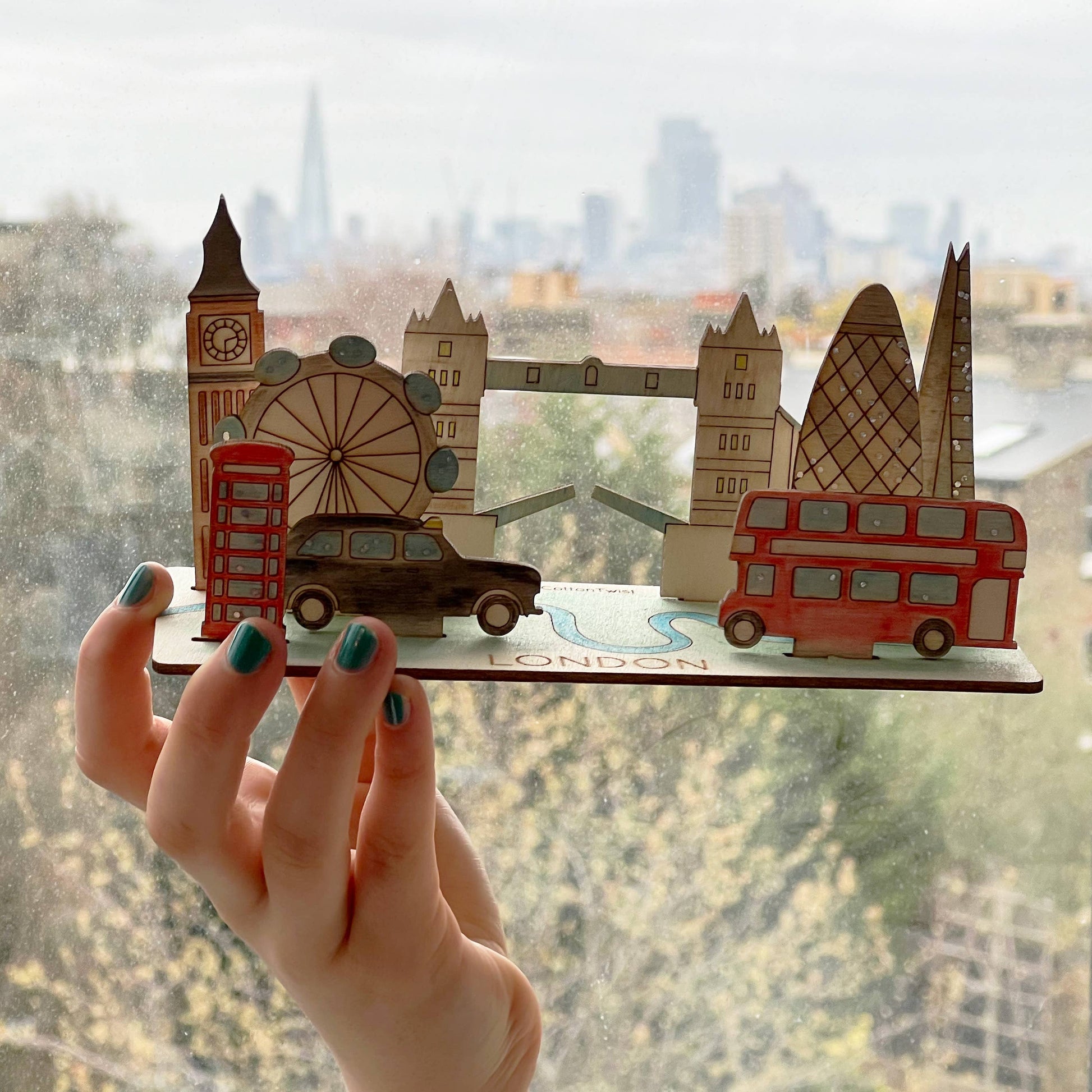 A hand holds up the fully assembled and hand-colored wooden 3D London model against a window, showing the finished scene in a natural light. The model features famous landmarks like the Tower Bridge, Big Ben, a double-decker bus, a taxi, and the London Eye, with a blurred city skyline visible in the background.
